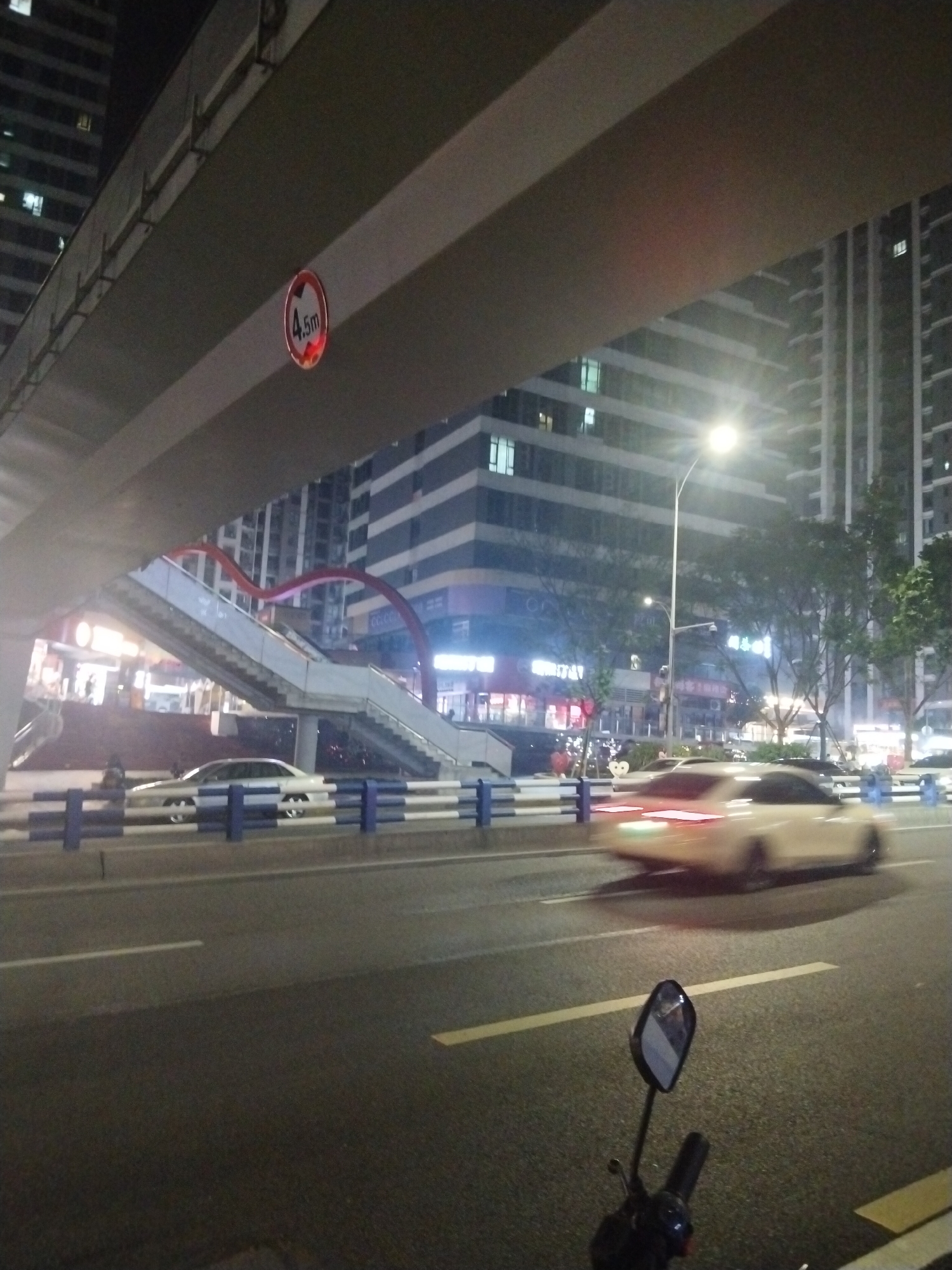 The image shows a street scene at night in the Jiulongpo District of Chongqing, China. A large overpass structure dominates the upper part of the frame, with a circular sign indicating a height restriction of 4.5 meters attached to its underside. Below the overpass, a pedestrian walkway bridge with red arches is visible, along with buildings. On the road, a white car is moving, creating a blur effect due to the motion. A motorcycle's mirror and handlebars are visible in the foreground. Street lights illuminate the area.
