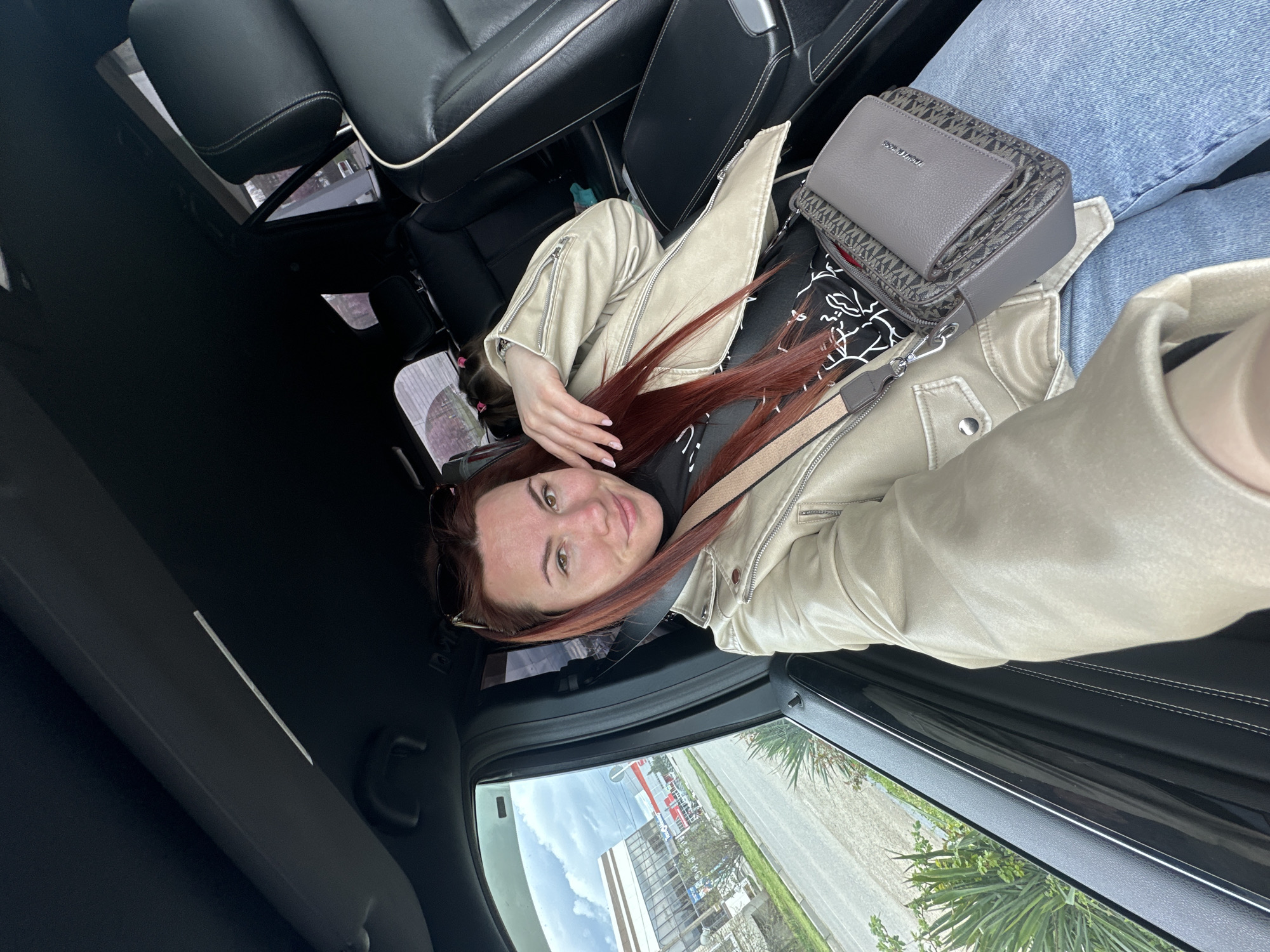 The image shows a woman with auburn hair lying in the passenger seat of a car. She is wearing a light-colored jacket and jeans, and a grey Michael Kors purse is positioned on her lap. The car is parked, and a view of Proletarskii, Russia can be seen through the window, showing buildings and greenery.