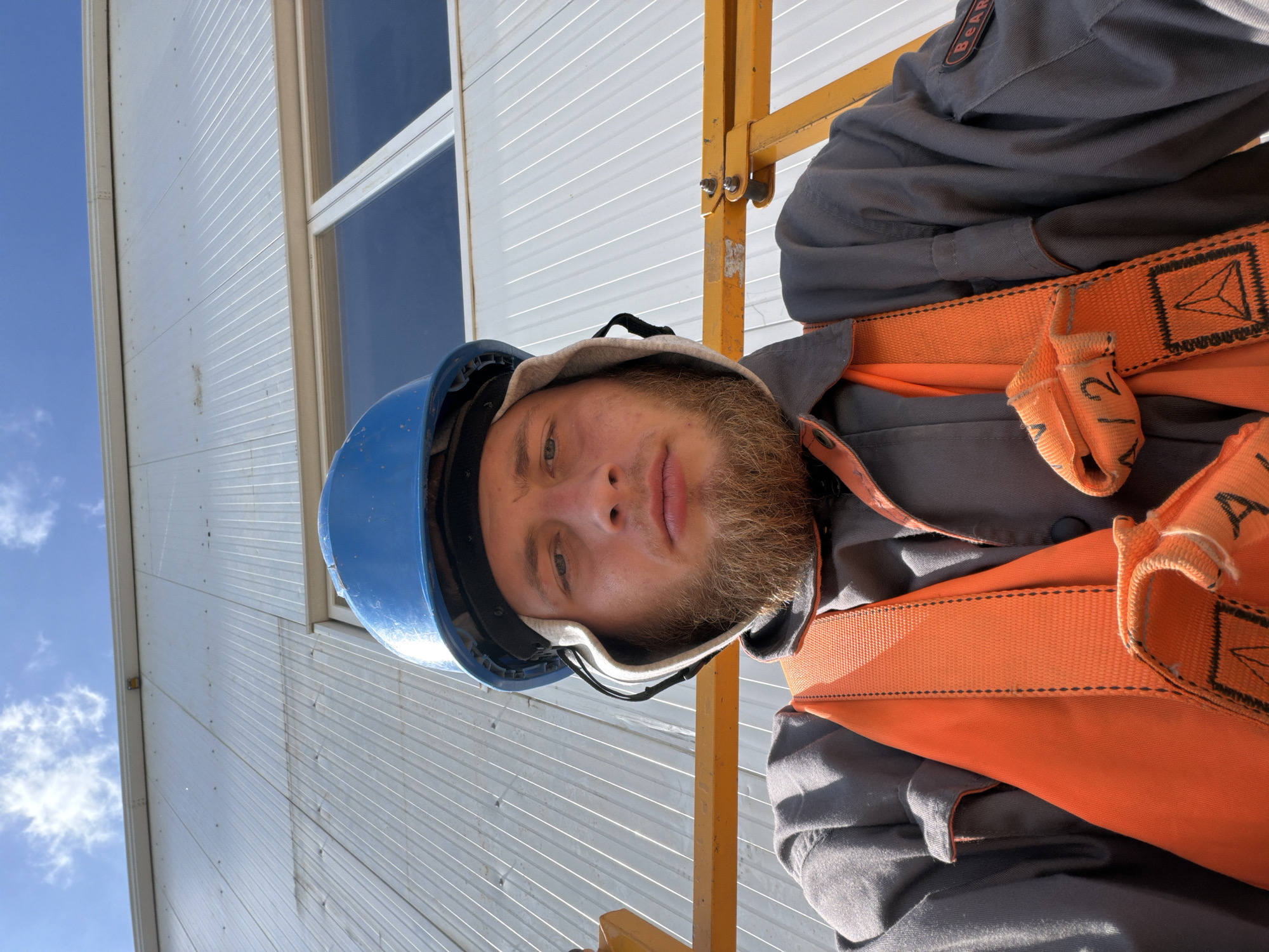 A man wearing a blue hard hat, grey work clothes, and an orange safety vest is taking a selfie. He has a beard and is looking at the camera. He is in front of a white, corrugated metal building with windows. The sky is blue with some clouds. The man appears to be on a yellow lift. The location is Wilkowisko, Poland.
