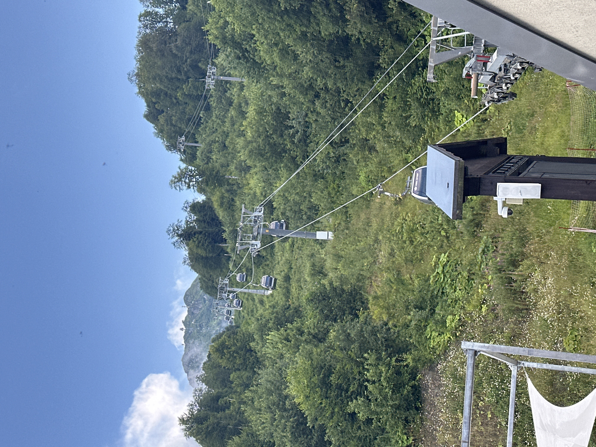 The image shows a mountain landscape with a ski lift and several cable cars ascending a lush green slope. The cable cars are dark and appear to be modern, with windows that allow a view of the interior. They are suspended from a thick cable that runs between tall support towers. The mountainside is covered in dense green trees and vegetation, with a clear blue sky overhead. In the distance, the peaks of higher mountains are visible, some shrouded in mist. The foreground shows part of a structure, possibly a station for the ski lift, with a security camera mounted on a wooden post. The overall scene depicts a picturesque natural setting, likely a popular tourist destination for outdoor recreation.
