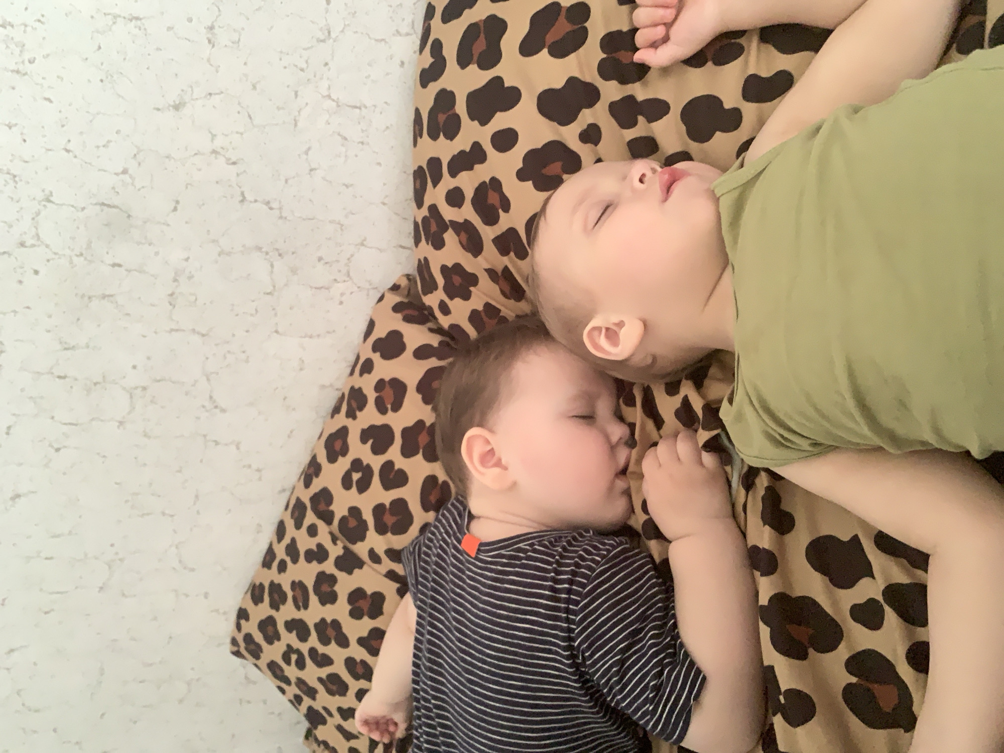 Two young children are captured from an overhead perspective, sleeping peacefully side-by-side on a leopard-print blanket or cushion. The child positioned higher in the frame, with fair skin and light hair, is wearing a plain light olive green short-sleeved top. Their eyes are closed, mouth slightly open, indicating deep sleep. Below this child, nestled closely, is another child, also with fair skin and light brown hair, sleeping soundly with eyes closed and hands gently curled. This child is wearing a dark blue or black and white striped short-sleeved top, with a small orange tag visible on the back of the collar.
The leopard print pattern features dark brown spots with lighter brown centers on a tan background, covering the area beneath both children. To the left and partially along the top and right edges of the image, a light, textured white surface is visible, likely a bedsheet, wall, or floor, providing a simple and clean background. The lighting is soft and even, suggesting an indoor setting during a nap time or night, conducive to sleep. The overall scene conveys a sense of calm and tranquility. There is no visible text in the image. The location context provided is Фоки, Russia.