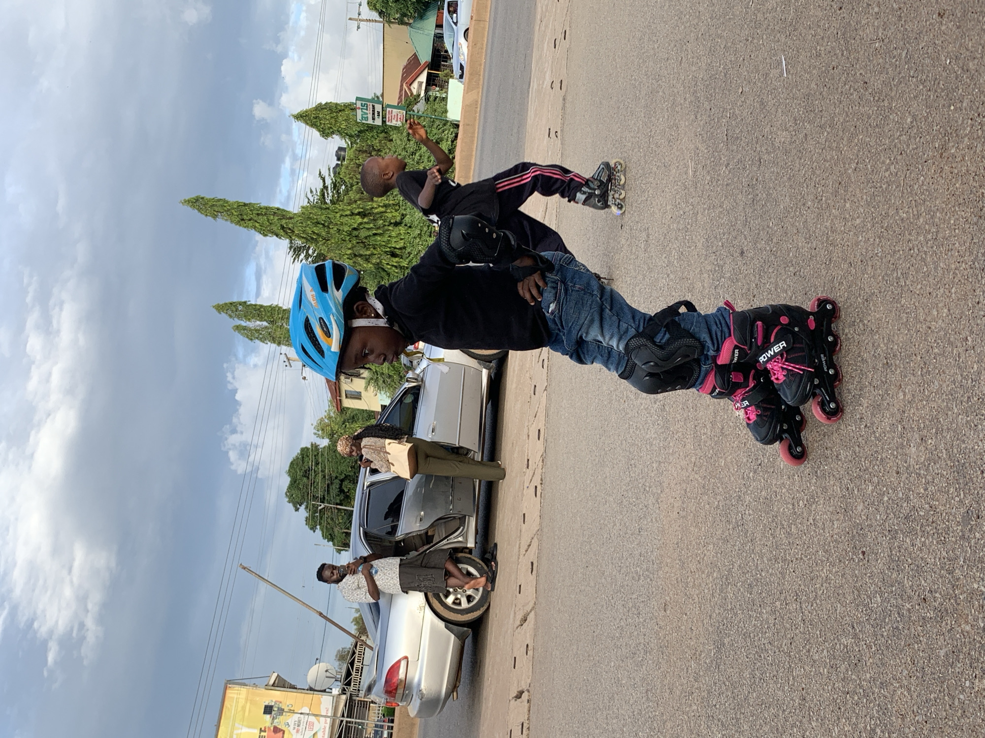 This image captures a roadside scene in Jos, Nigeria, distinguished by the "AVIS RESTAURANT & BAR" sign visible in the mid-ground right. The sky is largely overcast with white and gray clouds.
In the foreground, a child is depicted rollerblading on a paved asphalt road. This child, positioned to the left, wears a blue helmet with orange and white accents, a black long-sleeved top, blue jeans, black knee pads, and black rollerblades with pink wheels and laces, displaying "POWER" branding. The child's body is angled slightly, with hands on hips, suggesting a static or poised stance.
Further back and to the right, a second child is also rollerblading. This child wears a black t-shirt, dark pants with pink stripes down the sides, and black rollerblades. This child is actively in motion, leaning forward with arms slightly extended, moving from left to right across the frame.
In the mid-ground on the left, a silver four-door sedan is parked by the side of the road, with its rear passenger door open. An adult male, wearing a patterned short-sleeved shirt, dark shorts, and sandals, leans against the rear of the car while looking at a mobile phone held in his hands. Beside him, an adult female with a patterned head covering, a light-colored long-sleeved top, and olive-green trousers stands near the open car door, also engrossed in a mobile phone. A light-colored shoulder bag is visible on her left side.
The background features several tall, slender, coniferous trees, indicative of roadside landscaping. Electrical power lines traverse the upper portion of the frame. On the far left, a yellow building with a partially obscured sign is visible. The road surface exhibits some cracks and irregularities. Another white vehicle is partially visible moving in the far background on the right.