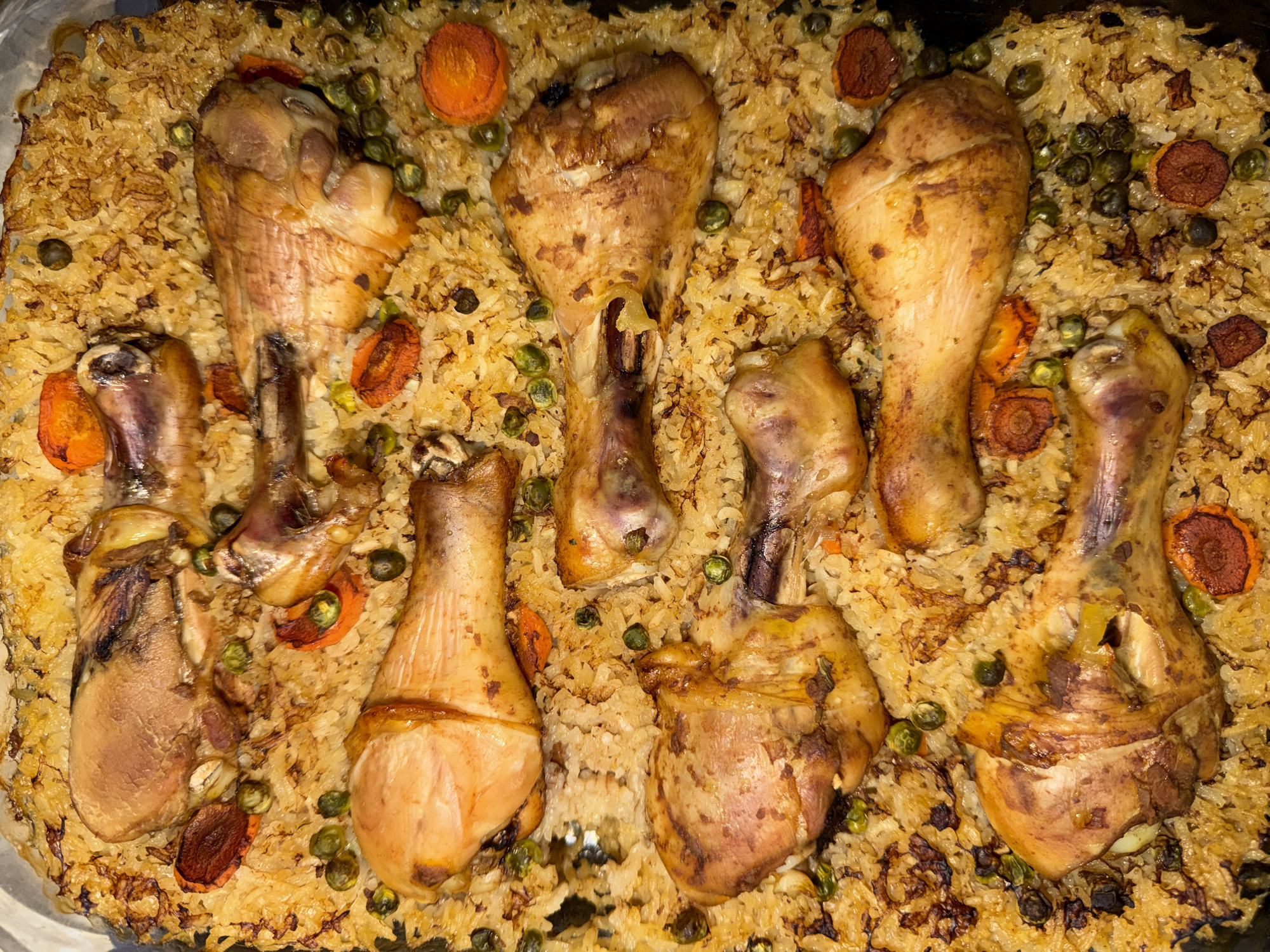 An overhead, close-up shot displays a large pan filled with a cooked chicken and rice dish, possibly a pilaf or paella. Eight chicken drumsticks or thighs, varying in size, are arranged within the golden-brown rice, which appears somewhat crispy and caramelized in parts, especially around the edges and beneath the chicken. The chicken pieces have a browned, roasted skin.
Scattered throughout the rice are numerous small, green peas and circular slices of cooked orange carrots, some of which also show signs of caramelization or roasting. The overall impression is a hearty, home-cooked meal presented in a communal serving dish.
There is no discernible activity taking place as the image captures a static food presentation. No people are visible. The lighting appears to be artificial, highlighting the textures and colors of the food. There are no visible location cues within the image itself, nor is any text visible. The time of day and weather cannot be determined.