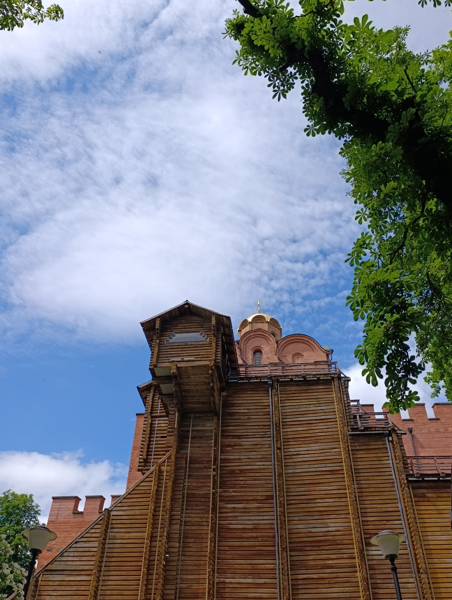 This image depicts the Golden Gate in Kyiv, Ukraine. The perspective is from below, looking up at the wooden and brick structure under a bright blue sky with scattered white clouds. Lush green leaves from a tree frame the top right corner of the image, casting some shade. The Golden Gate itself is constructed with a prominent wooden facade and a brick upper section topped with a golden dome. Architectural details include multiple windows and balconies. In the foreground, a lamppost is visible to the left. The scene appears to be a clear day with good visibility.