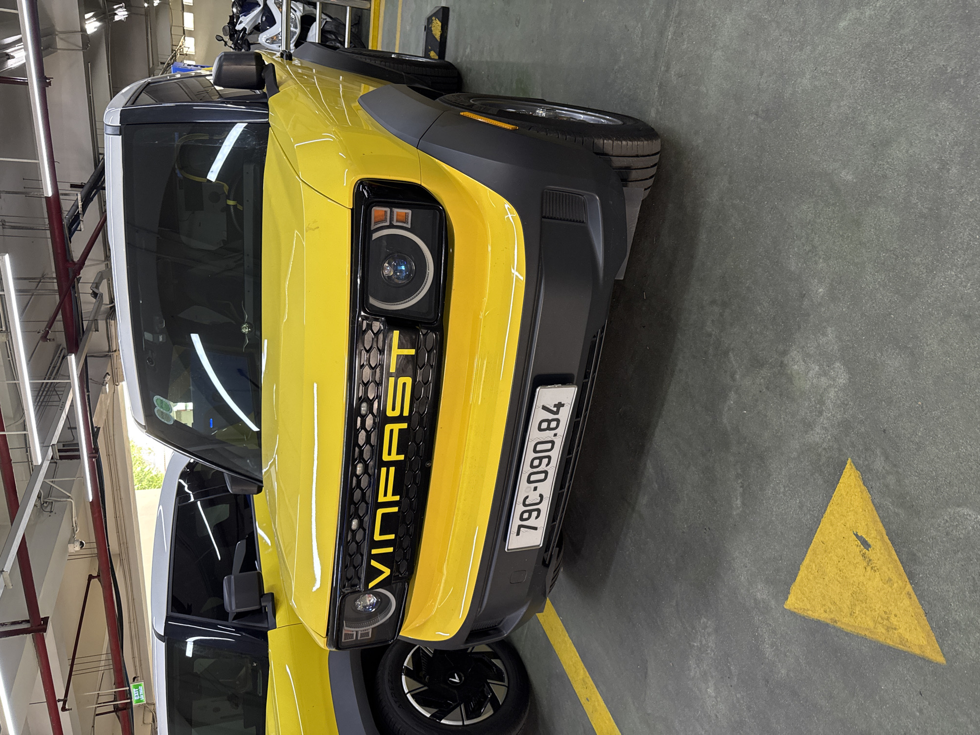 The image shows the front of a bright yellow VinFast electric car parked indoors, likely in a covered parking garage or showroom in Nha Trang, Vietnam. The car has a black grille with the VinFast logo in yellow letters. The headlights are square with circular daytime running lights. A license plate with the characters "79C-090.84" is visible on the front bumper. To the left of the main car, another yellow VinFast car is partially visible, also with a white roof. The floor is concrete with yellow painted lines, including a triangular marker. In the background, metal pipes and lighting fixtures are visible on the ceiling, along with a portion of a white motorcycle. The environment appears to be an industrial or commercial indoor space.