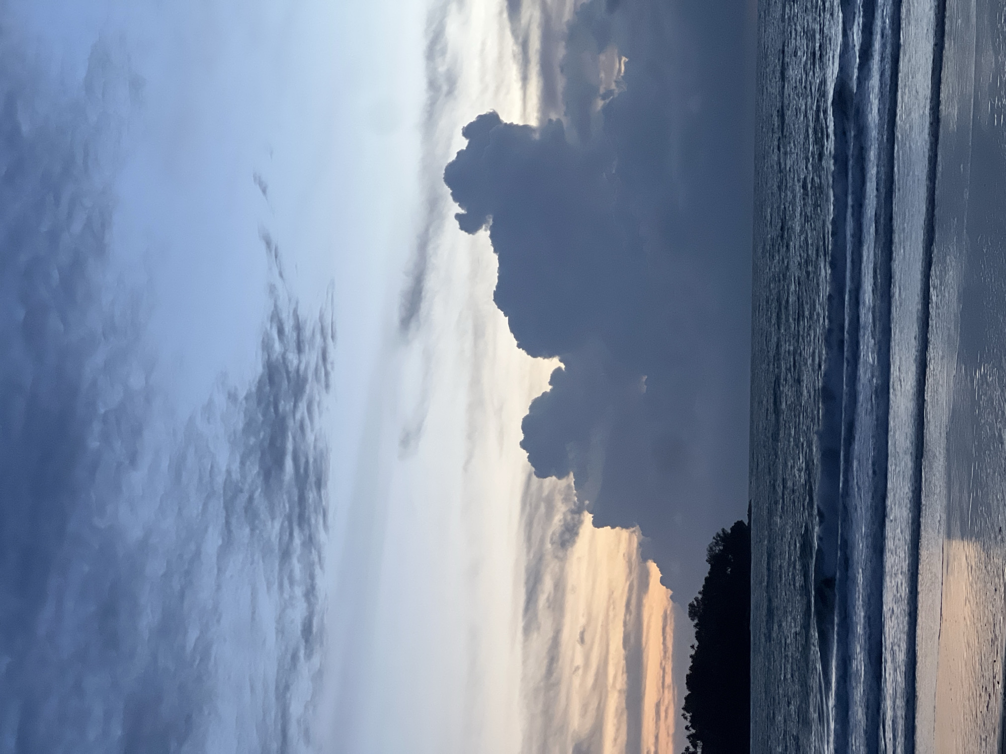 A seascape captures the twilight sky over the ocean at Choeng Thale, Thailand. The sky displays a gradient of colors, with soft oranges and yellows near the horizon, transitioning to blues and grays higher up, dotted with scattered clouds. A large, dark cloud formation dominates the center of the sky, appearing almost sculptural. To the left, a dark silhouette of a tree-lined headland juts into the water. The ocean is calm, with gentle waves rolling towards a sandy beach, reflecting the soft light of the sky. The water is a deep blue-gray, with streaks of white foam from the breaking waves. The beach in the foreground is wet and reflective, showing subtle colors from the sky. No people or animals are visible in the scene. The overall atmosphere is serene and tranquil, typical of a peaceful evening by the sea.