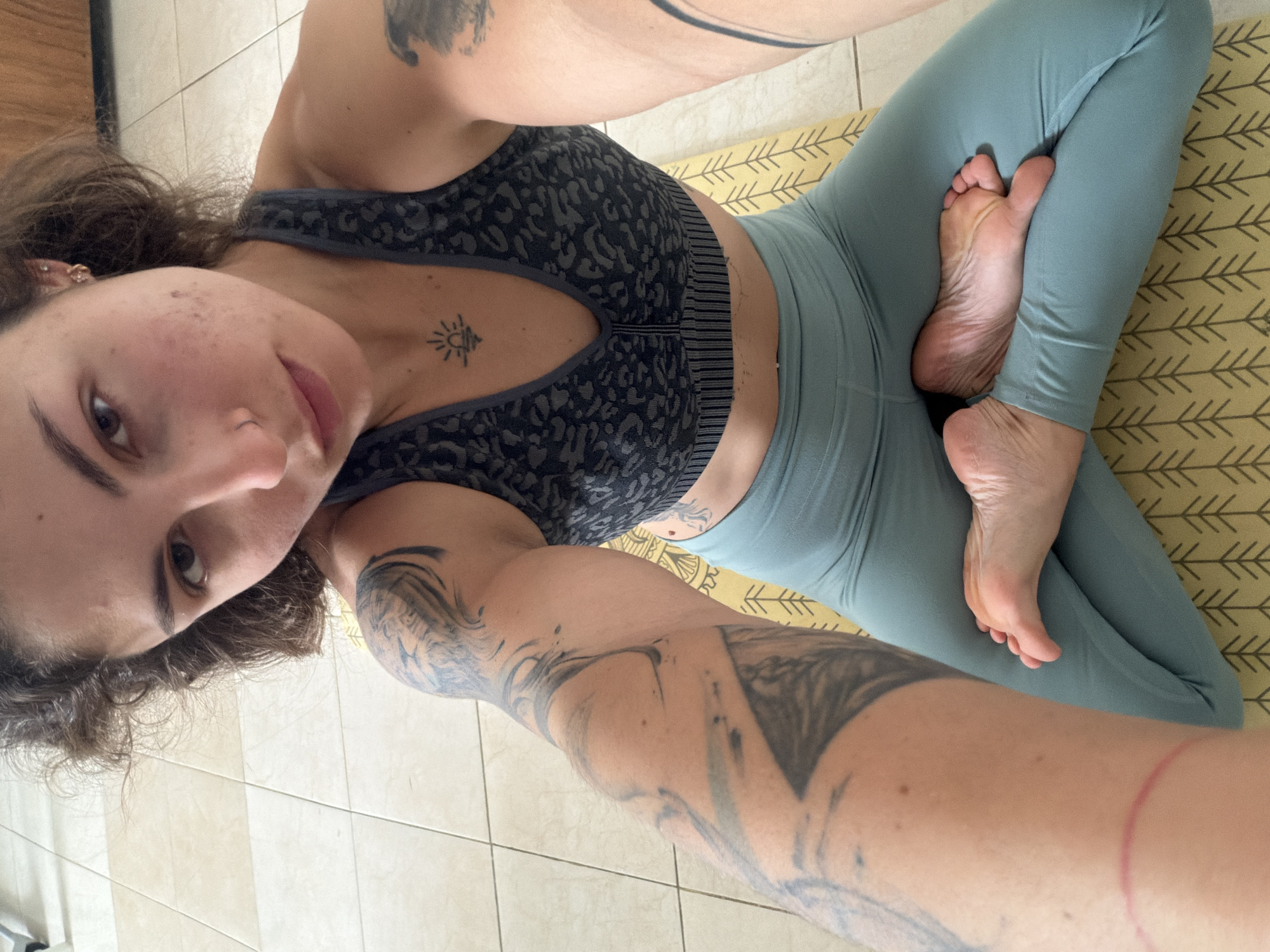 A woman with brown curly hair and visible tattoos is lying on her back on a yellow patterned yoga mat. She's wearing a dark gray sports bra with a patterned design and light blue leggings. Her legs are crossed in a yoga position, with one foot resting on the opposite thigh. The location is likely a residence in Kiryat Bialik, Israel, as indicated by the tiled flooring. The lighting suggests it is daytime. The woman is taking a selfie.