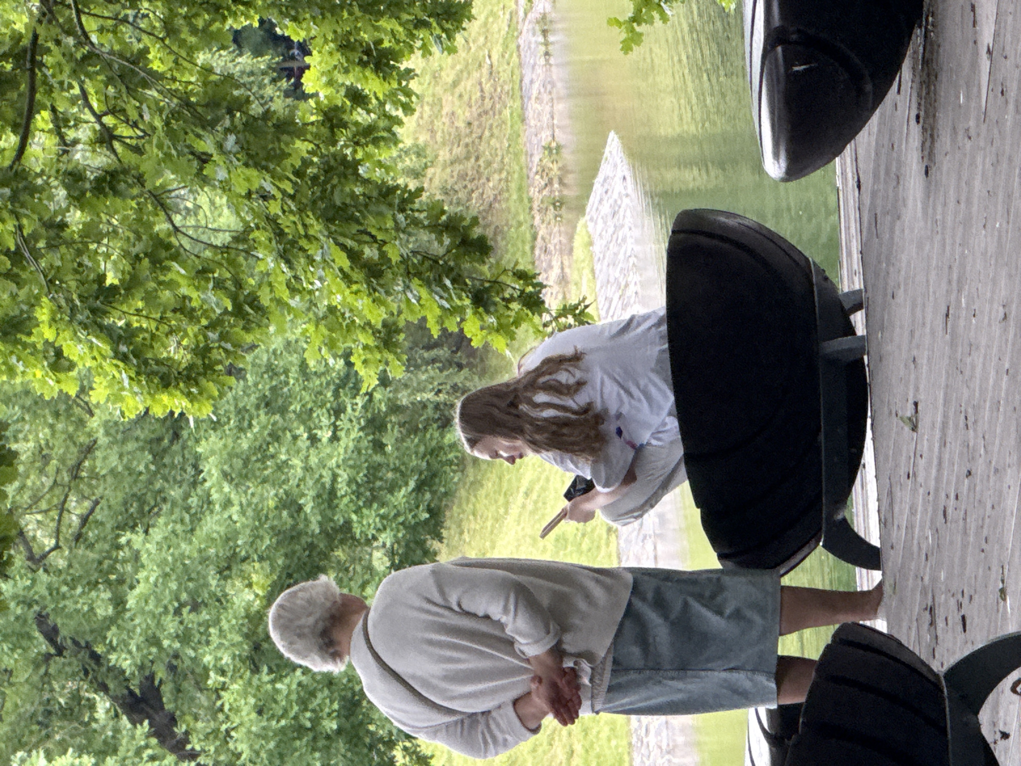 An outdoor scene shows two people and a body of water. On the left, an older person with grey hair stands with their back to the camera, wearing a light-colored sweater and a denim skirt. Their hands are clasped behind their back. To the right, a younger person with long brown hair crouches on a wooden platform, facing away from the older person and towards the water. They are wearing a light-colored top with some purple and pink design on the back and light-colored pants. The younger person is holding a smartphone. In the foreground, two black, curved benches or seating structures are visible on the wooden platform. The background consists of a grassy hill and lush green trees, with a calm body of water reflecting the greenery. The scene appears to be a park or waterfront area.