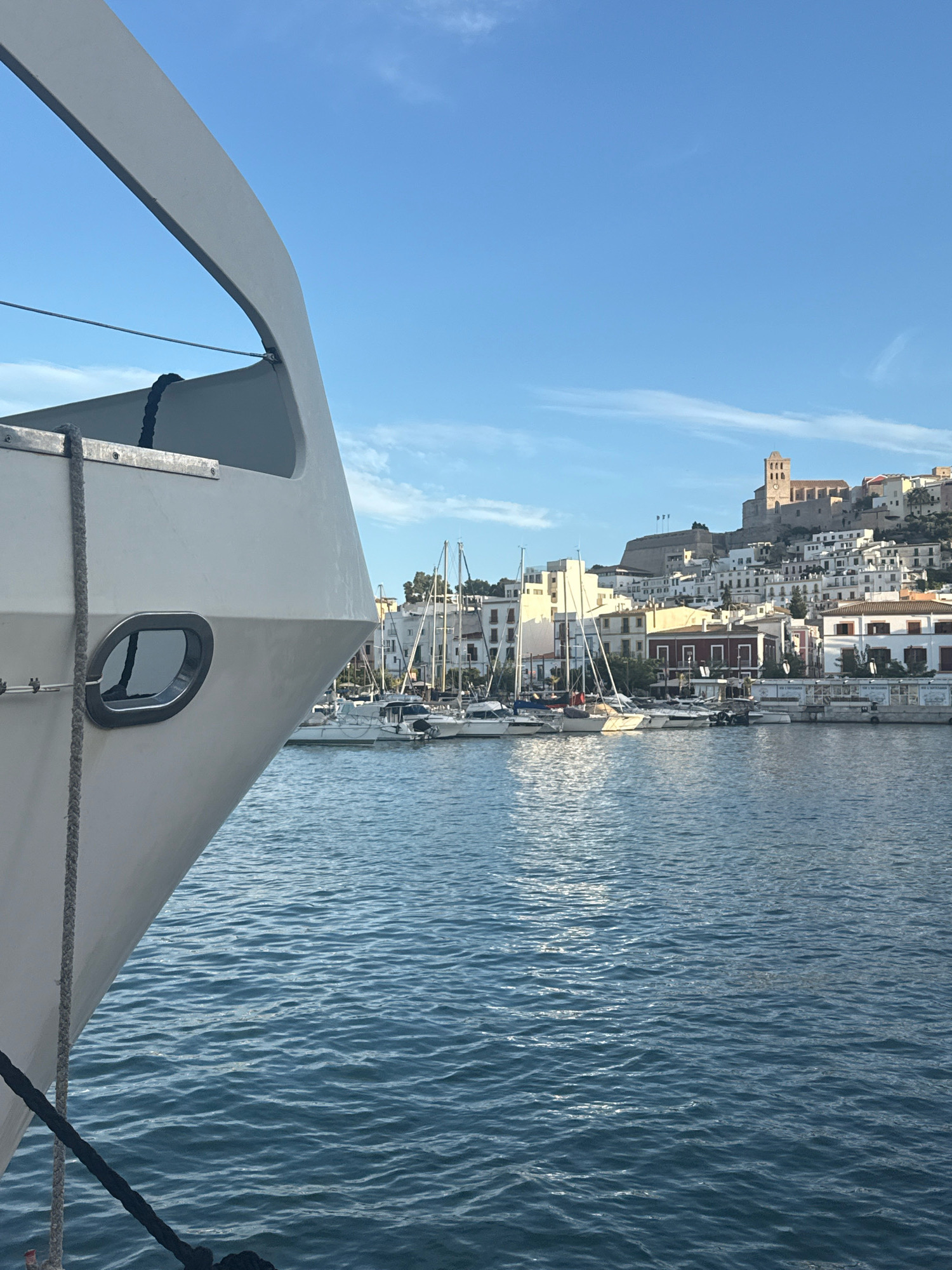 An eye-level shot captures a portion of a white yacht's bow in the foreground, facing right, with a dark rope secured to it and leading out of frame. Beyond the yacht, a body of blue water stretches across the frame, rippling gently. In the background, a white cityscape climbs a hill under a clear blue sky with scattered wispy clouds. Numerous sailboats with tall masts are docked along the waterfront. The cityscape is dominated by white buildings with terracotta roofs, and a prominent fortress-like structure with a bell tower crowns the hill. The scene is brightly lit, suggesting it is daytime. The overall impression is one of a sunny day in a European coastal city. This is likely the Port of Ibiza, with the Dalt Vila (Old Town) and Ibiza Cathedral visible on the hill.