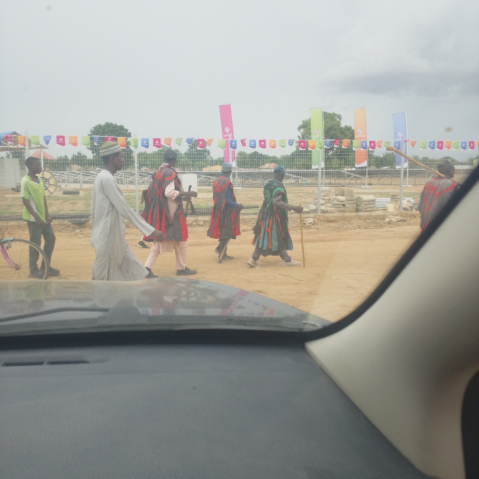 The image captures a group of individuals walking across a dirt path in Wuro Ngangandi, Nigeria. In the foreground, the interior of a vehicle is visible, with part of the dashboard and windshield frame. A young man wearing a green t-shirt and dark pants stands to the left, with a bicycle with a pink frame beside him. To his right, a man in a light-colored garment and a patterned cap walks away from the camera. Further to the right, three other men are walking in the same direction. Two of these men are wearing traditional attire consisting of brightly colored cloaks with red and black accents over light-colored pants. One of these men carries a long stick. The background features a fence and construction materials, with several tall, colorful flags and a string of smaller flags overhead. Trees and buildings are visible in the distance under a cloudy sky. The scene appears to be outdoors in a semi-open area.