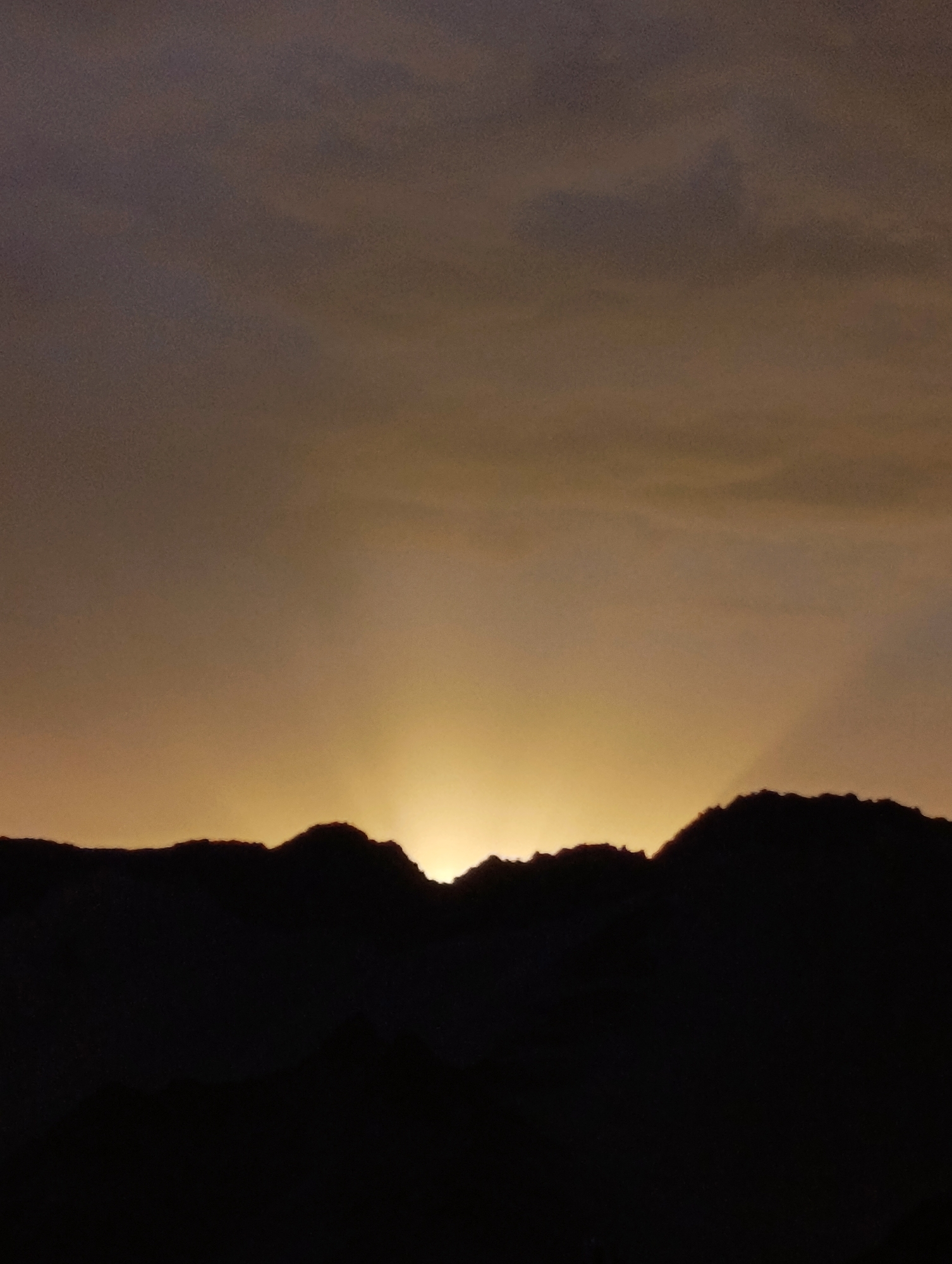 The image shows a mountain range silhouetted against a dusky sky. The sky is a gradient of dark gray at the top transitioning to a warm, golden hue near the horizon, where the sun is either rising or setting. The mountain peaks are dark and jagged, with a few points of light peeking through the gaps between them, suggesting the sun is low on the horizon. The foreground is entirely in shadow, obscuring any details of the ground or vegetation. The scene evokes a sense of tranquility and the vastness of nature. While the image is beautiful, it does not provide any specific visual cues or landmarks that would allow for the identification of its location within Leh, India.