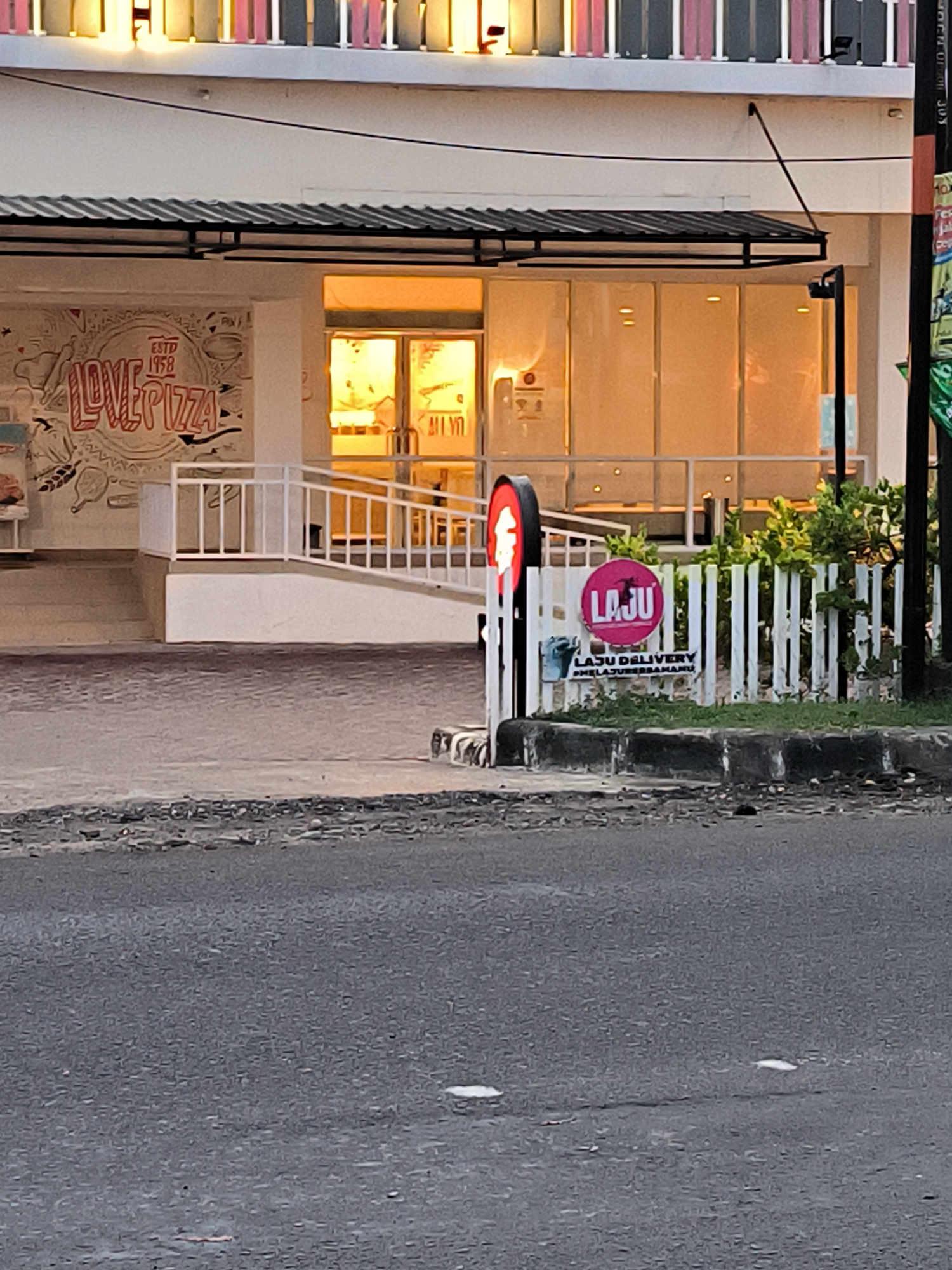 This image captures a street-level view of a commercial building, likely a restaurant, in Lhokseumawe, Indonesia, during what appears to be the late afternoon or early evening. The setting is urban, with an asphalt road in the foreground and a paved area leading up to the building.
The main subject is a brightly lit building with a white facade. A black corrugated metal awning extends over the entrance area. To the left, a large, stylized mural on the wall reads "LOVE PIZZA" with "ESTD 1958" above it, indicating a pizza establishment. The interior of the building, visible through large glass panels, is bathed in a warm, golden light, suggesting indoor lighting is on.
An accessibility ramp with white railings leads up to the building's entrance. On the right side of the image, beyond a low curb, there is a small patch of green foliage and a white picket fence. Attached to this fence are two signs. One is a circular pink sign with "LAJU" written in white, and below it, "LAJU DELIVERY" and "#MELAJUKANUSAMAMU" are visible. To the left of this fence, a red and black circular sign, possibly a traffic or business sign, is partially obscured. No people or specific activities are visible in the frame, giving the scene a quiet, still quality. The overhead structure of the building features decorative elements in pink, grey, and white.