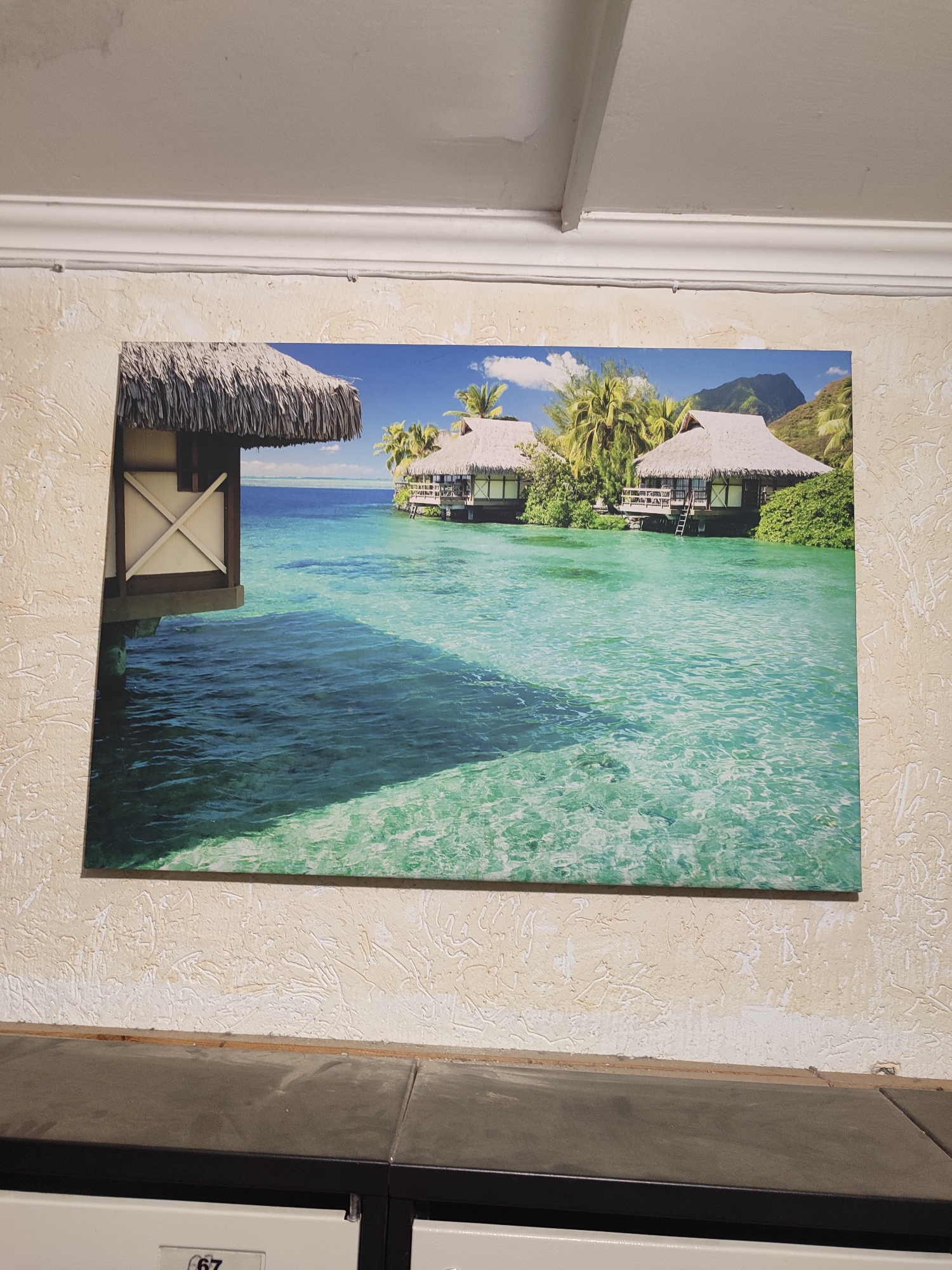 An interior overhead shot captures a large rectangular print of a tropical overwater bungalow scene, affixed to a textured, light beige wall. The print depicts several thatched-roof bungalows situated over calm, turquoise water, with lush green palm trees and distant mountains under a blue sky with scattered white clouds. To the left, a portion of one bungalow's side is visible, featuring a brown frame and an "X" shaped brace. The wall itself exhibits a rough, stucco-like texture with irregular white markings and appears to be in an indoor setting. Above the print, a white, decorative ceiling molding separates the wall from a smooth, off-white ceiling. A thin, grey cable runs horizontally along the wall just beneath the molding. Below the print, the upper surface of a dark-colored, possibly laminate or stone, piece of furniture is visible, likely a countertop or cabinet top. Below this, a lighter-colored metal panel, resembling a locker or drawer, is partially visible with the number "67" printed on it. No people are visible in the scene.