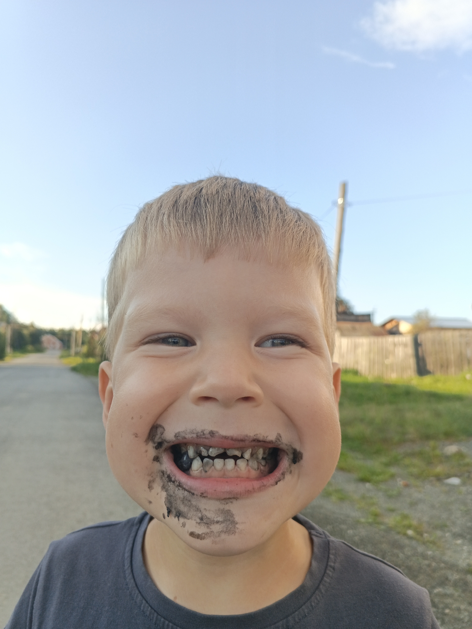 A young boy with blond hair is pictured from the chest up, looking directly at the camera with a wide, dirty smile. His mouth is open, revealing dirty teeth and some dirt smeared around his lips and chin. He is wearing a dark gray t-shirt. The background is blurred, showing a road and some houses with wooden fences. The sky is clear blue with a few wispy clouds. The setting appears to be outdoors, possibly in a rural or suburban area. The location is identified as Medvedyovka, Russia.