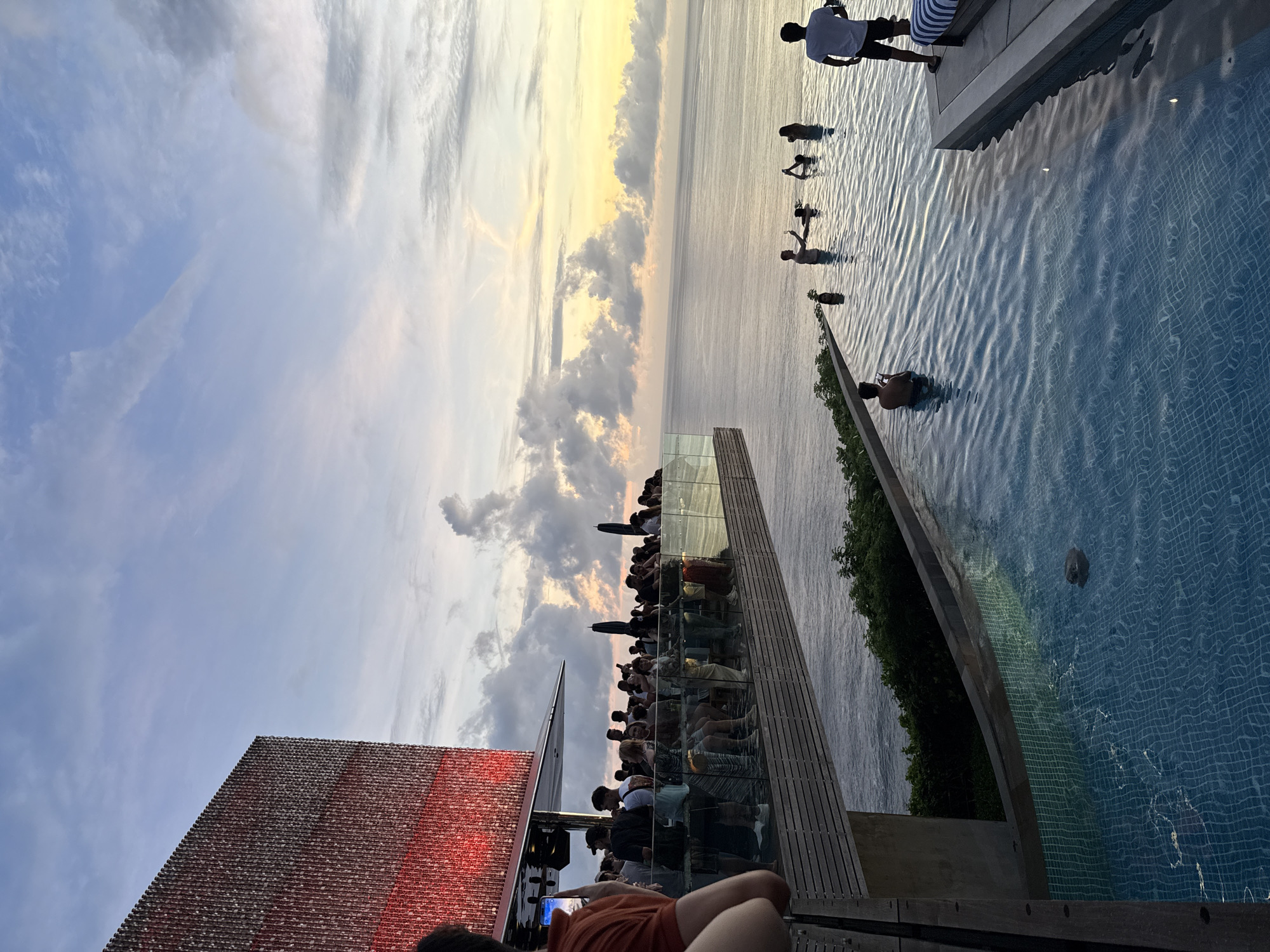 This image shows a scene at an establishment overlooking the ocean in Kuta Selatan, Indonesia. The view appears to be from a rooftop or elevated platform. A large group of people are gathered behind a glass barrier, seemingly enjoying the sunset or ocean view. To the right, an infinity pool stretches out, with several individuals swimming or standing in the water. A person in a white shirt and dark shorts stands by the edge of the pool, while others are submerged or partially visible. The pool tiles have a blue grid pattern. To the left, part of a building structure is visible, with an unusual texture and design on its roof or facade. The sky is filled with clouds, and the sun is setting, casting a warm glow on the horizon. The overall atmosphere suggests a lively, tourist-oriented location.