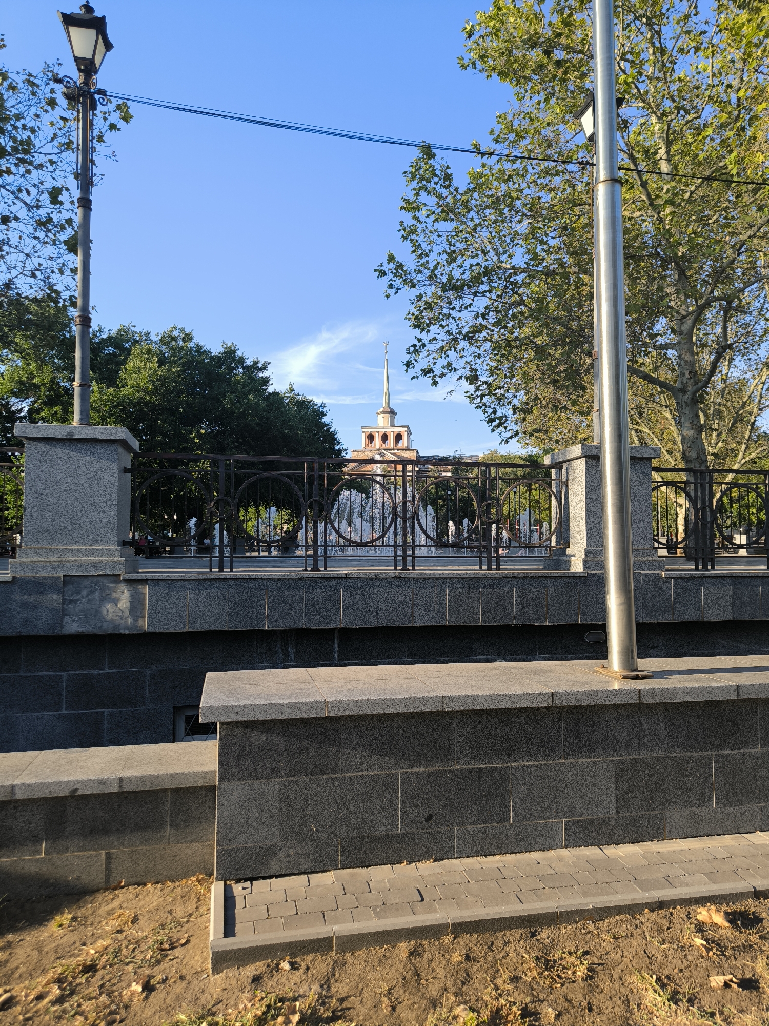 This image depicts a sunlit scene in Mykolaiv, Ukraine, likely a public space with a fountain and park elements. In the foreground, a low stone wall and a paved walkway are visible. Above this, a granite-clad structure forms the base of a railing with a dark metal fence featuring circular motifs. Behind the fence, a fountain with multiple jets of water is active, with a prominent building featuring a spire visible in the background, surrounded by green trees. A streetlamp with an old-fashioned design stands on the left side of the frame, and a modern, polished metal pole is positioned on the right. The sky is clear and blue, indicating a pleasant day. There are no people clearly visible in the frame.