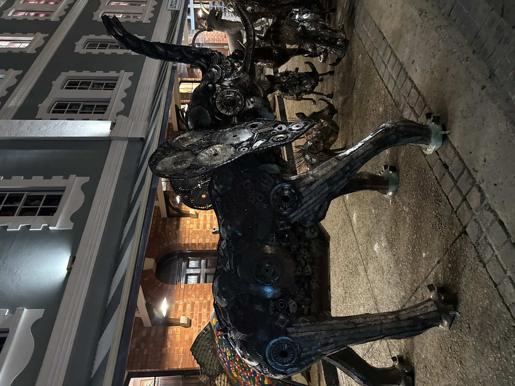 The image shows a metal sculpture of an animal, possibly a bull or a fantastical creature, located on a cobblestone-paved area beside a building. The sculpture is made of various metal parts, including gears, chains, and other mechanical components. The building in the background has a distinct architectural style with ornate window frames and a grayish-white facade. Adjacent to this building is a brick structure with an arched doorway, illuminated by warm lighting. The scene appears to be at night, based on the lighting conditions. The location is Cape Town, South Africa.