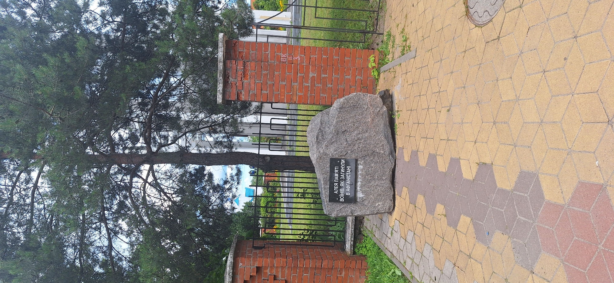 The image depicts a memorial stone located in the city of Bila Tserkva, Ukraine. The stone, with an inscribed plaque, is situated on a paved walkway made of hexagonal and rhombus-shaped bricks in yellow, gray, and red. Two brick pillars, one on the left and one on the right, frame the scene, with a black metal fence behind the memorial stone. In the background, a large pine tree stands tall, its branches spreading across the frame. Beyond the fence, glimpses of buildings and greenery can be seen, including a building with a blue roof. The sky is partly cloudy. The plaque on the stone reads, "Алея пам'яті воїнів, які загинули в Афганістані" which translates to "Alley of Memory of Soldiers Who Died in Afghanistan." The scene appears to be a public space, likely a park or a memorial site.