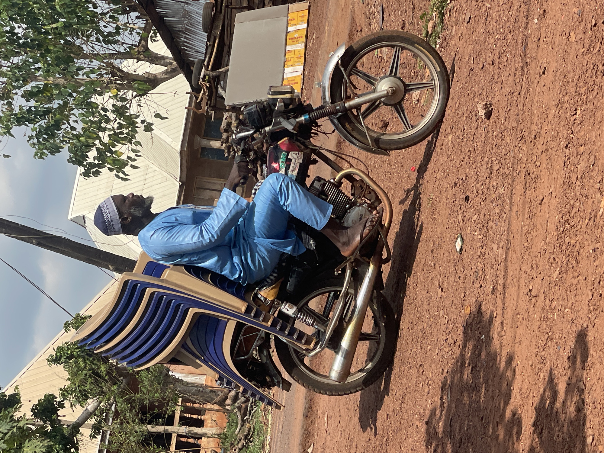 A man is depicted seated on a motorcycle heavily loaded with stacked chairs, paused on a reddish-brown dirt road in Ogbe, Nigeria. The image is taken during bright daylight, casting distinct shadows on the ground.
The primary subject is a middle-aged or older man with dark skin and a graying beard, looking towards the left of the frame. He is dressed in a light blue traditional tunic and a blue and white patterned cap. He sits on a motorcycle that has been adapted to carry a substantial load. Strapped securely to the rear of the motorcycle is a tall stack of approximately eight plastic chairs, featuring blue seats and beige frames. The motorcycle appears utilitarian, with a visible engine (where "MG" can be seen on a part), an exhaust pipe, and a front wheel with a five-spoke design.
In the background, behind the motorcycle and the man, there are simple buildings with corrugated metal roofs. Some trees with green foliage are also visible, suggesting a mix of natural and built environments. To the right, near one of the buildings, cardboard boxes are stacked, with partial text possibly reading "OREBEA". The ground is a dry, textured dirt road with small stones, characteristic of many unpaved areas. The sky is bright with some light clouds. The scene captures a common mode of transport and commerce in the region.