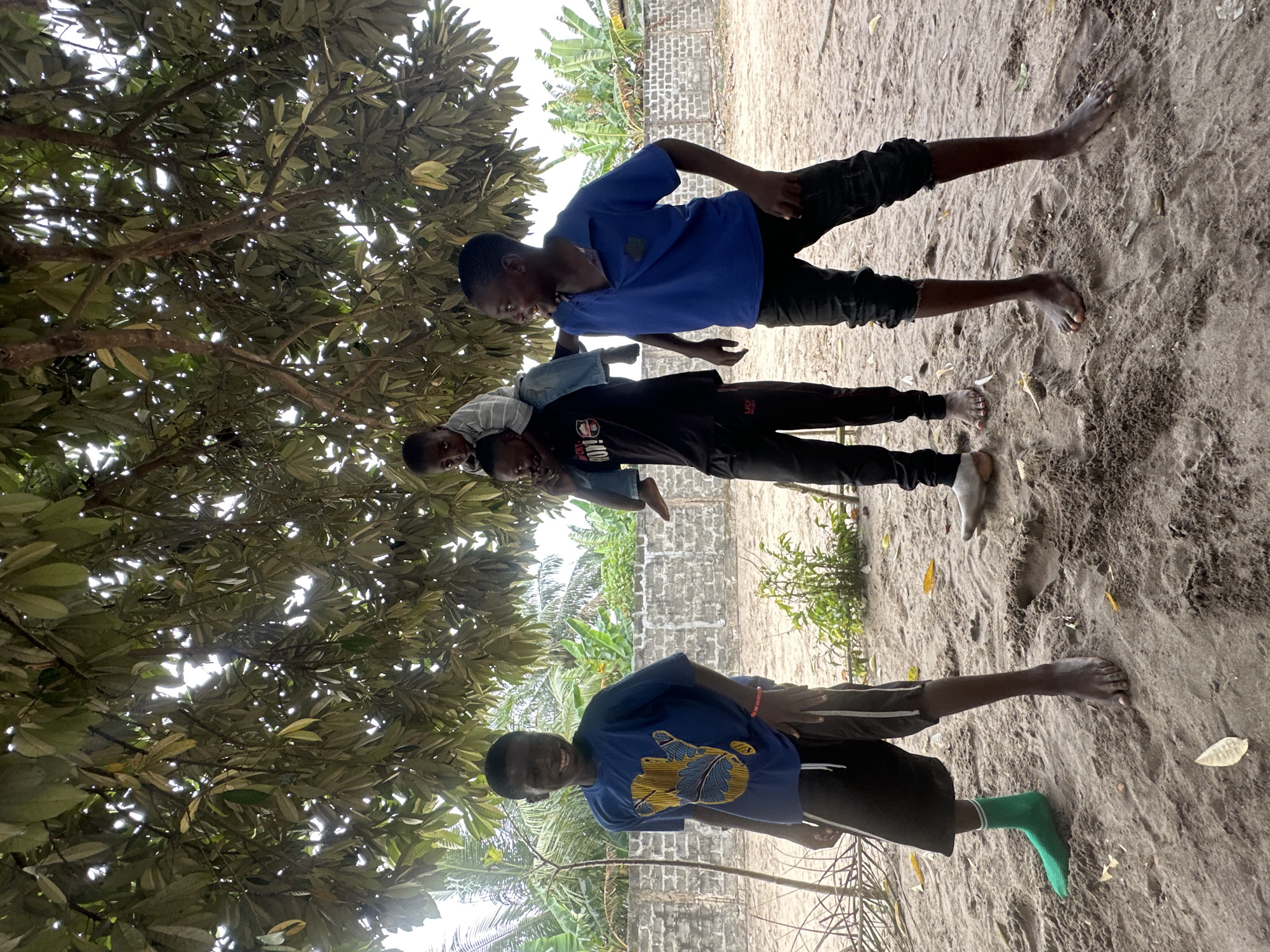Three young Black boys are standing under a large tree with dense foliage in an outdoor, sandy area. The boy on the left, wearing a blue t-shirt with a yellow and blue design and dark shorts, smiles directly at the camera, with one green sock and one bare foot visible. In the center, another boy in a black t-shirt and dark pants has a younger boy on his shoulders; the boy on the shoulders wears a striped long-sleeved shirt and jeans. The third boy, on the right, is wearing a bright blue polo shirt and dark pants, looking to his right with an open mouth as if speaking or laughing. A low, grey brick wall with vegetation behind it forms the backdrop, suggesting a location in the city of Badagry, Nigeria, where such walls and tropical plants are common. The ground is sandy with scattered leaves.