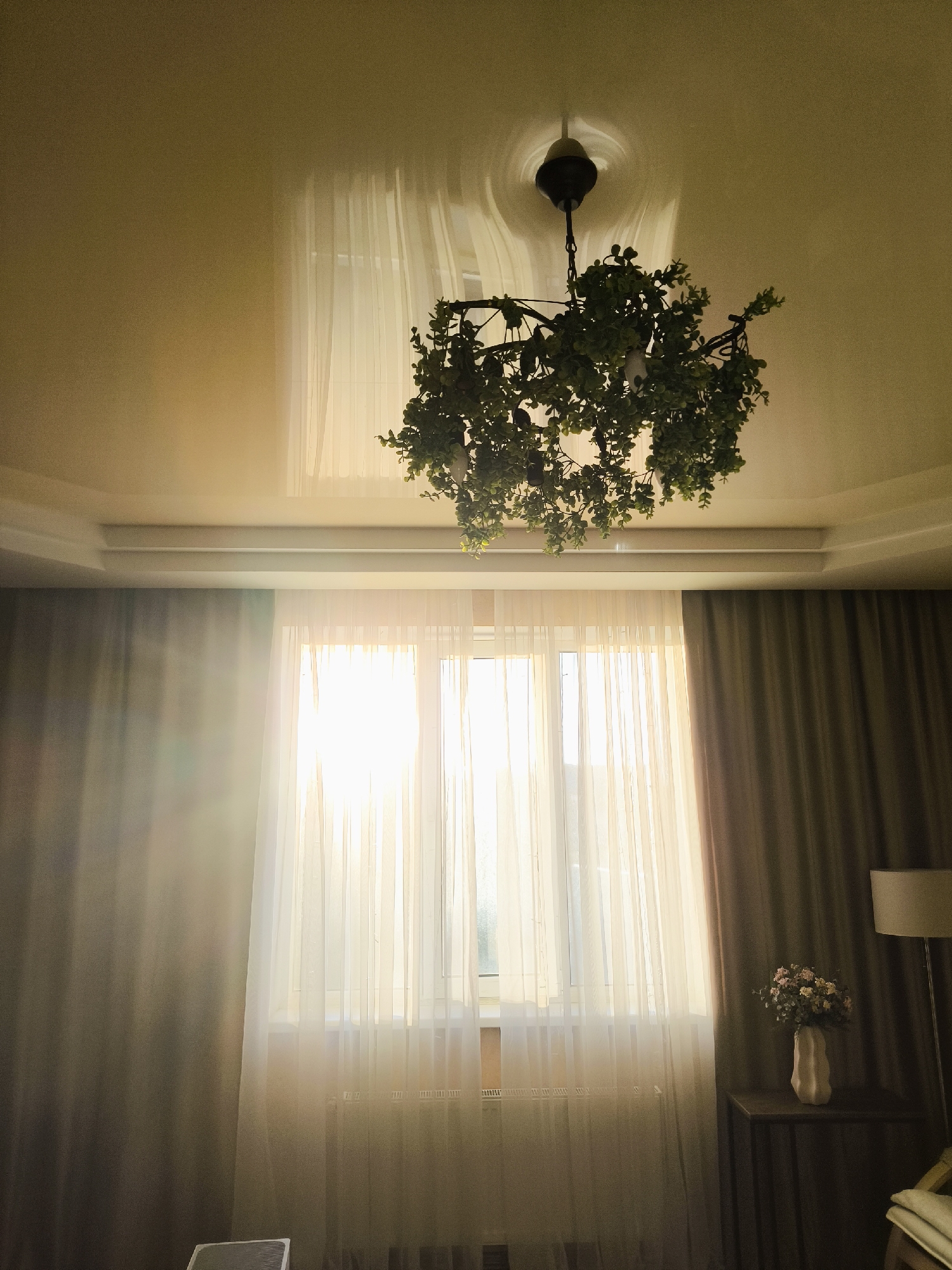 The image depicts an indoor scene, likely a living room or bedroom. A chandelier adorned with artificial greenery hangs from the ceiling, casting a circular light reflection on the textured ceiling. The room is illuminated by natural light streaming through a large window covered by sheer white curtains. To the right of the window, dark brown curtains are drawn. On a small dark wooden table next to the window, a vase containing a bouquet of pink and white flowers is visible, with a floor lamp to its right. The walls appear to be painted a light color, and there is a radiator beneath the window. The floor is not visible in the frame.