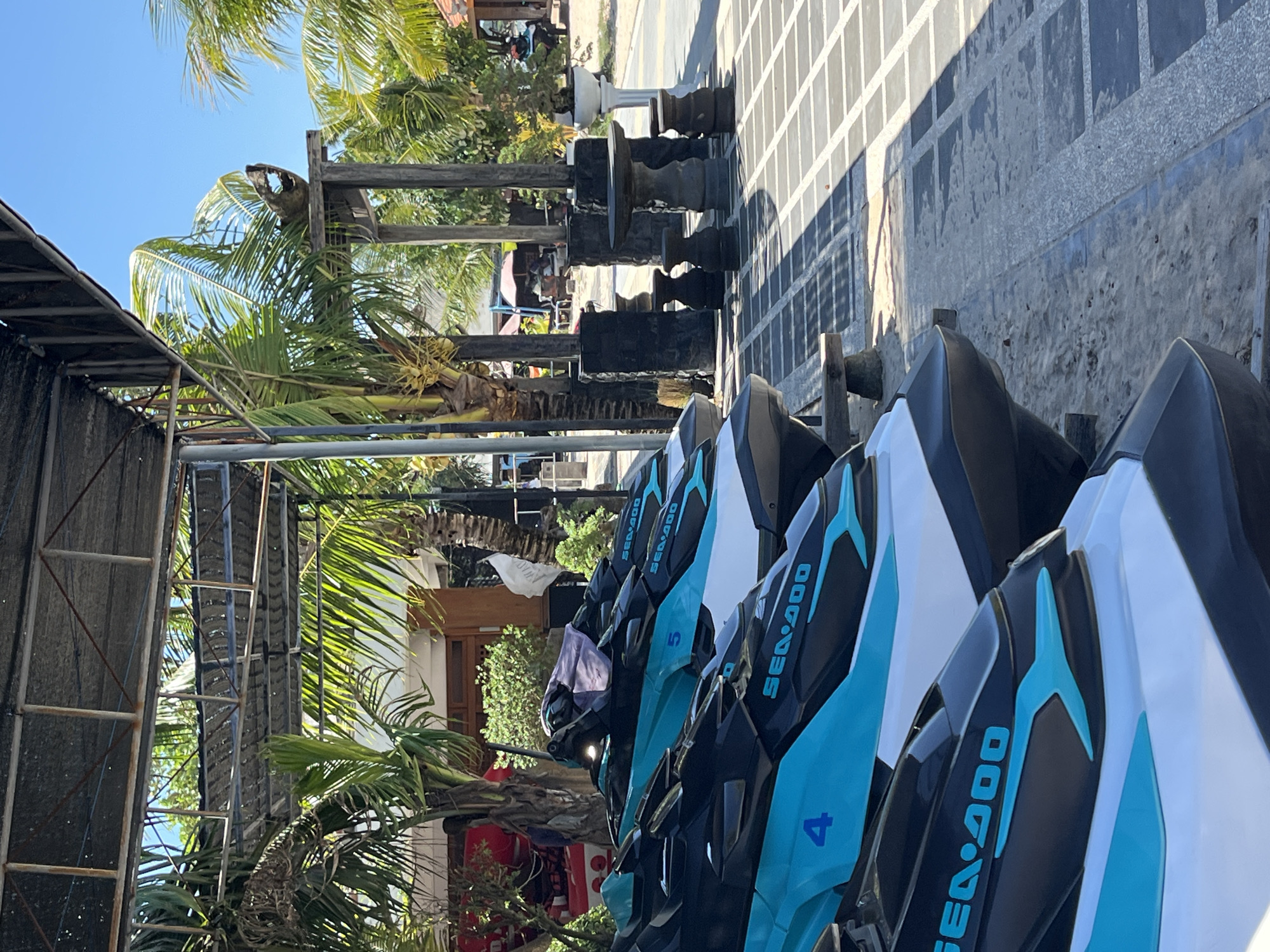The image depicts a row of jet skis on display in an outdoor setting, possibly a rental or sales location. The jet skis are branded "SEA-DOO" and numbered "4" and "5," with a color scheme of black, white, and teal. They are parked on what appears to be a sandy or concrete surface, adjacent to a sheltered area with metal framing and what looks like a metal roof. In the background, there are palm trees and other vegetation typical of a tropical climate. Further back, there are structures including what may be a house or shop. On the side are decorations or furniture. The overall impression is of a tourist or recreational area in a coastal or island location, possibly in Indonesia.