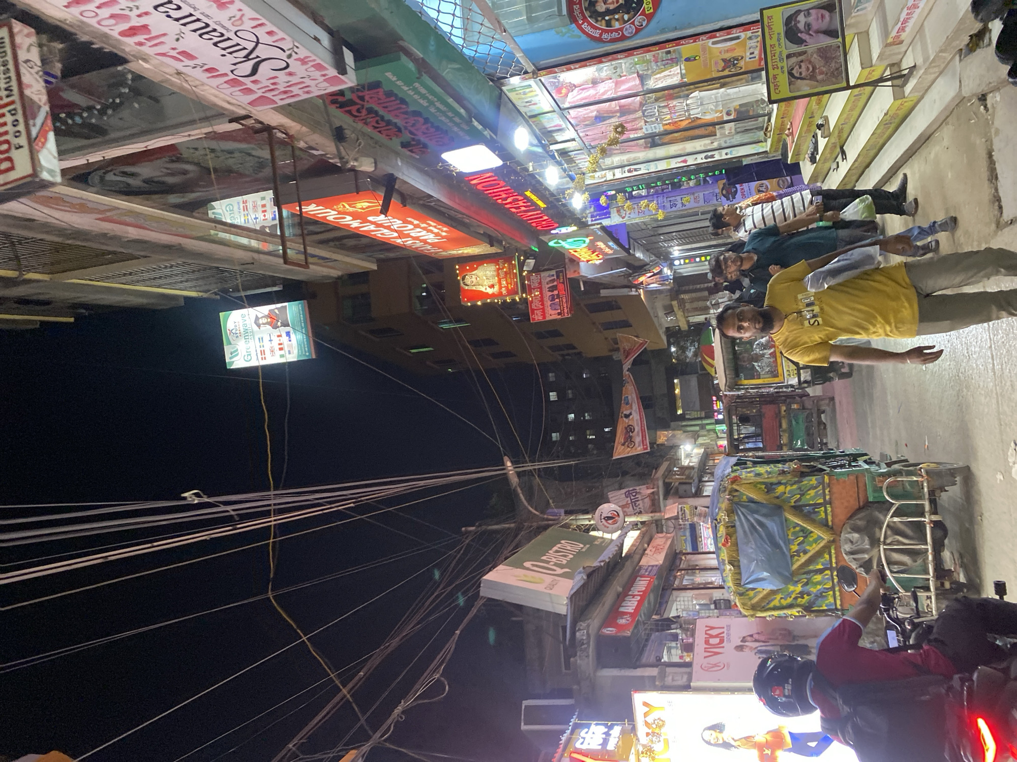 This is a photograph taken at night on a crowded street in Dhaka, Bangladesh. The street is lined with shops, their signs illuminated by neon lights. Above the shops, a dense network of electrical wires crisscrosses the sky. In the foreground, a person on a motorbike wearing a red shirt, helmet, and backpack is seen. Adjacent to this person, there is a camouflaged rickshaw. Several pedestrians are walking on the right side of the image. Multiple signs of various businesses, including "Q-BISTRO," "Skinaura," and "Greenwave," are visible. The scene is typical of commercial areas in Dhaka, characterized by high density and vibrant street life.
