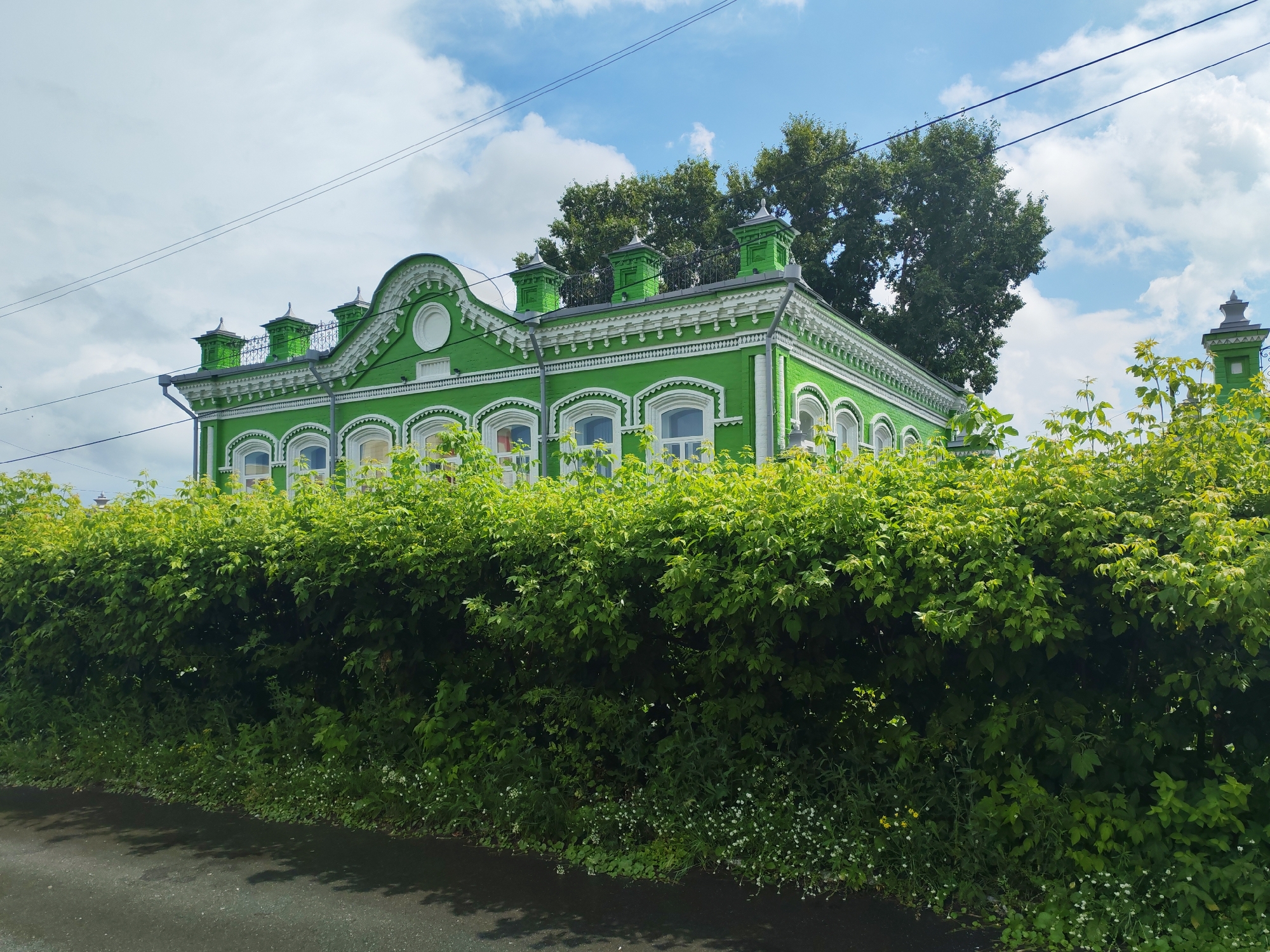 A bright green, two-story building with white trim and decorative eaves sits behind a dense, tall green hedge. The building has arched windows on the first floor and smaller, rectangular windows on the second. The roof is peaked with several decorative dormer-like structures. Trees are visible behind the building. The foreground shows a paved surface, possibly a road, with shadows cast on it from the foliage. The sky above is blue with scattered white clouds. The scene appears to be in a residential or historical district of an unspecified city.