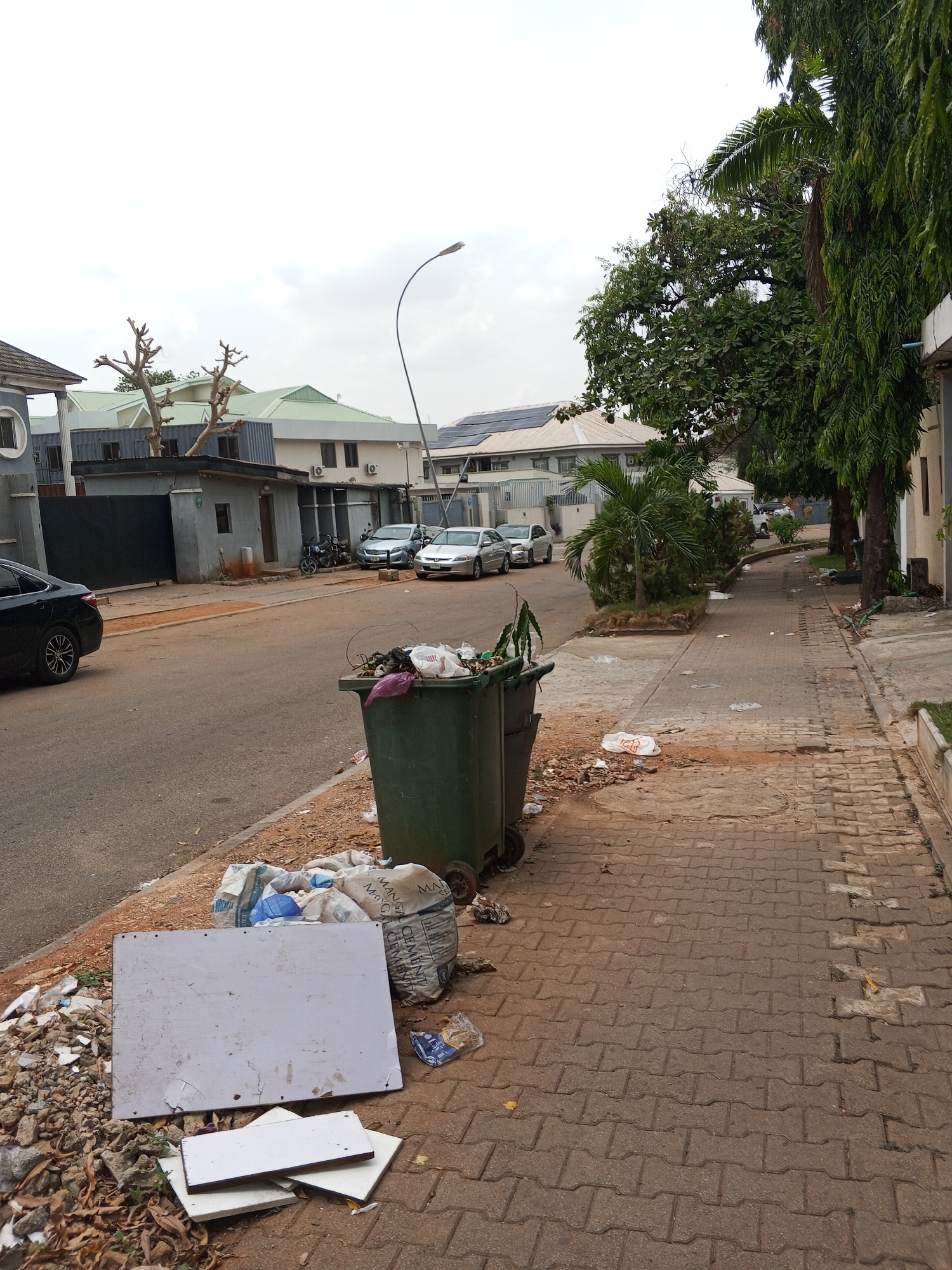 The image shows a street scene in Abuja, Nigeria. The road is paved and leads towards several buildings with light-colored roofs, some equipped with solar panels. Cars are parked along the street. A black vehicle is partially visible on the left side of the frame.
On the right, there's a paved sidewalk bordered by trees and some greenery. A significant amount of garbage is scattered along the sidewalk and overflowing from two green trash bins located near the road. Debris includes plastic bags, building materials, and paper. There are two bags of what appears to be cement. One of the boards lying on the sidewalk is white and rectangular.
The scene generally suggests a lack of proper waste management in the area. A curved streetlight is present in the background.