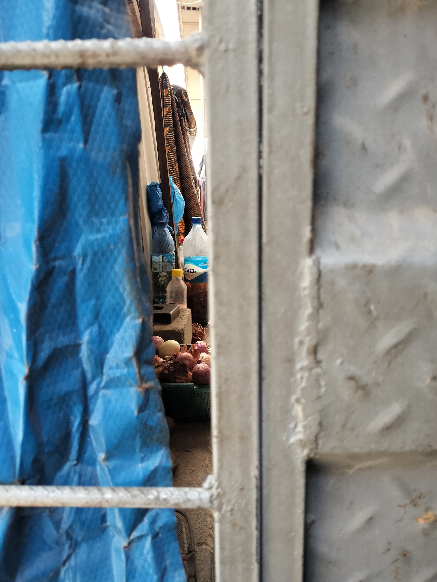 Through a grey metal gate and a blue tarp, the scene reveals an informal vendor's setup. There's a basket filled with onions, bottles of water (including one brand-labeled "eva"), and a small yellow bottle. The vendor's items rest on concrete blocks. In the background, fabric hangs vertically, suggesting wares for sale. The rough texture of the metal gate and tarp, coupled with the basic arrangement of goods, is consistent with the informal economy commonly found in Lagos, Nigeria. No people are visible in the frame.