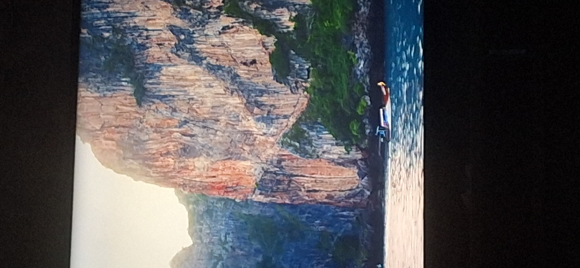 The image shows a scenic view of dramatic, towering cliffs covered in lush green vegetation along a coastline. The cliffs have a textured, stratified appearance with varying shades of orange, brown, and gray. Sunlight illuminates the upper portions of the cliffs, creating highlights and shadows. In the foreground, the deep blue water of the sea is visible, with subtle ripples on its surface. A long, narrow boat with a blue and white canopy and a tan stern is navigating through the water near the base of the cliffs. The boat appears to be a traditional longtail boat, commonly found in Southeast Asia. The overall scene evokes a sense of natural grandeur and tranquility. There is no discernible connection to the city of Bila Tserkva, Ukraine, as the landscape and boat are characteristic of a tropical or subtropical coastal region, not Eastern Europe.