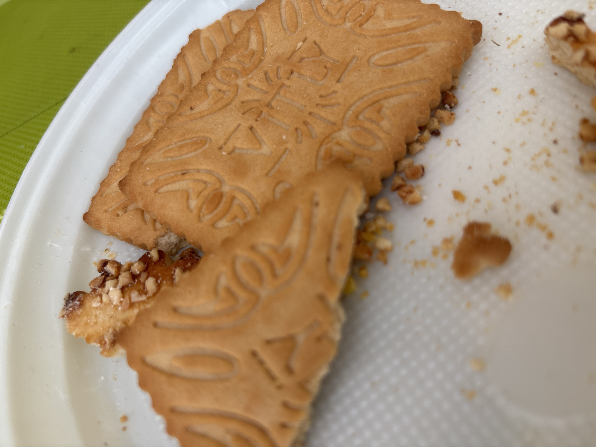 The image shows several patterned rectangular cookies on a white plate. Some have been partially eaten, revealing a filling with nut pieces. Crumbled cookie pieces are scattered around the plate. The setting appears to be outdoors, with a glimpse of a green surface visible in the upper left corner of the image. There is no verifiable information to suggest any connection to the city of Верхнеказымский, Russia.