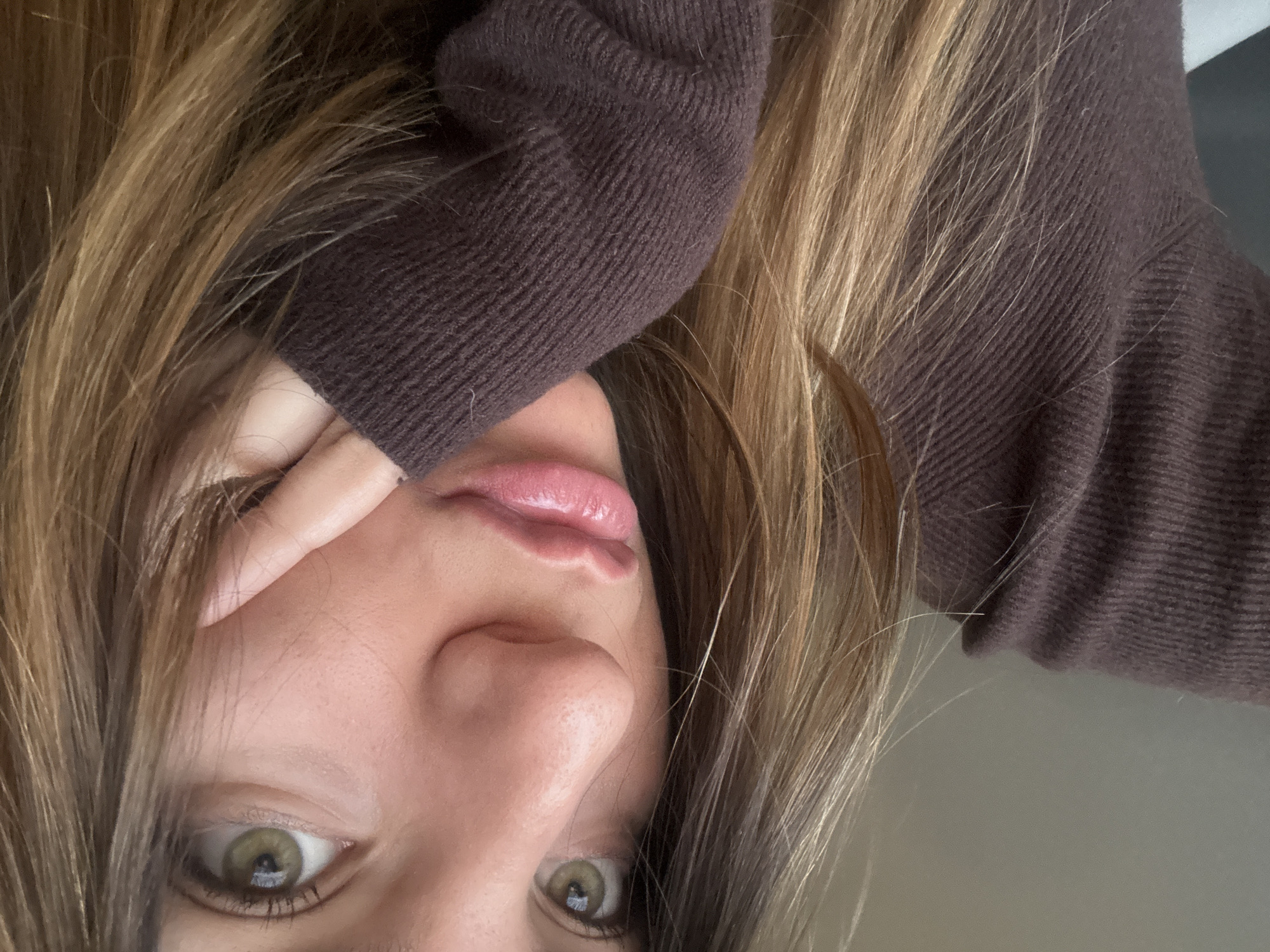 A close-up, angled view of a person's face. The person has light brown hair with blonde highlights and green eyes. They are wearing a brown sweater and their fingers are partially covering one eye. The background is out of focus and appears to be a light gray wall. The location is Unknown, Unknown.