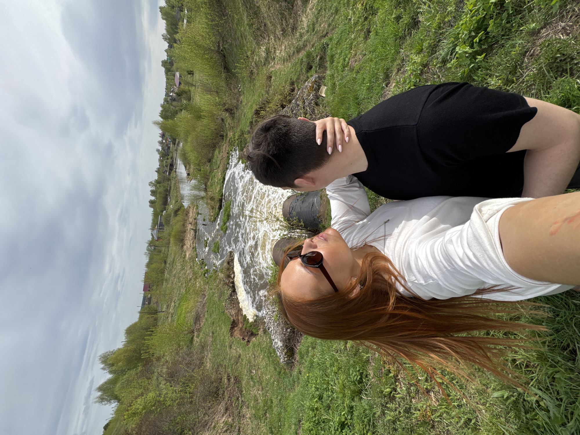 Two people are next to a small waterfall. A person in a black shirt has a hand on the neck of a person in a white shirt and sunglasses, and the person in the white shirt is looking up at the person in the black shirt. They are in Nozhovka, Russia.