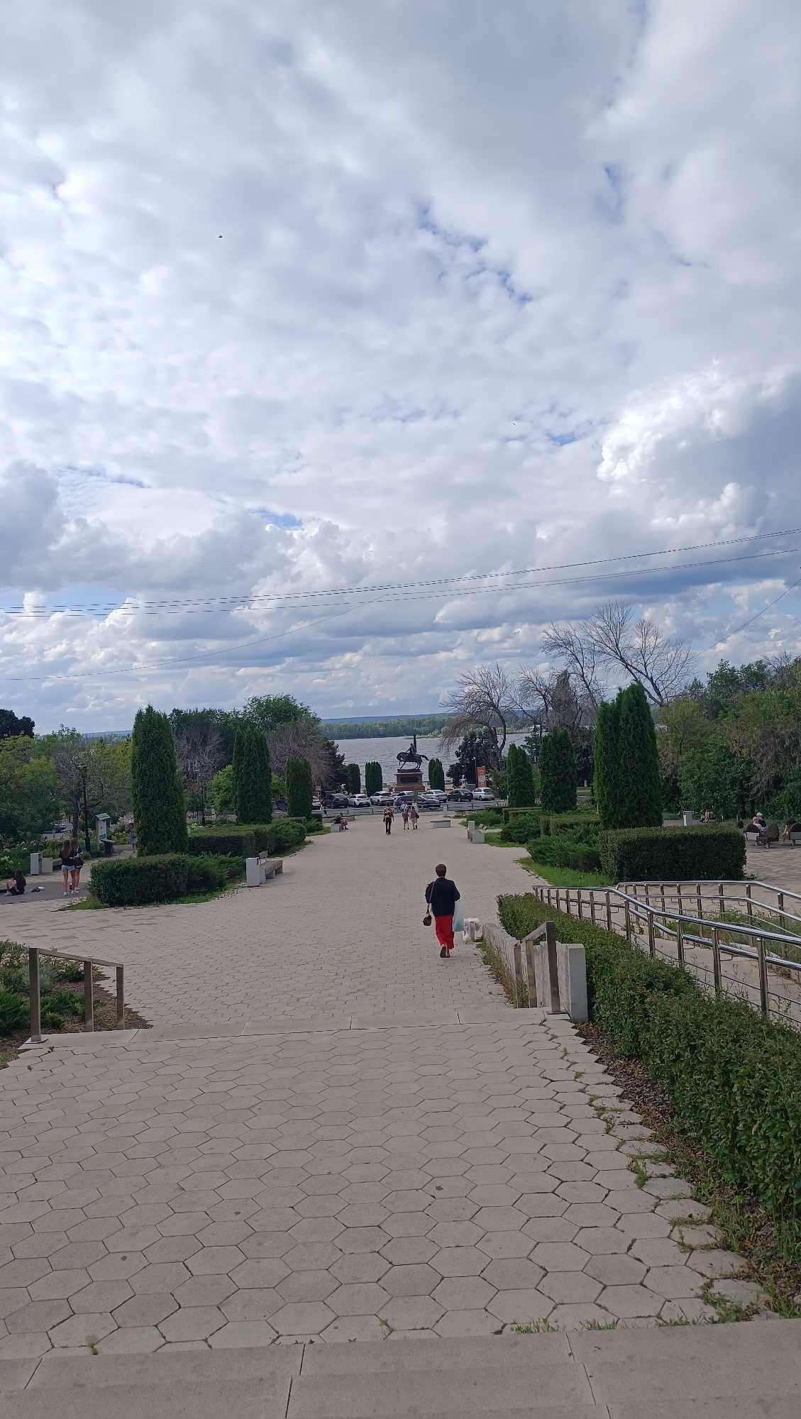 The image depicts a paved walkway leading down a gentle slope in a park, likely located in Samara, Russia, given the context. The walkway is made of hexagonal concrete tiles and is flanked by manicured hedges and tall, conical evergreen trees. In the distance, at the end of the walkway, a statue of a man on horseback is visible, situated near a body of water, which appears to be the Volga River. The background shows a hazy sky with scattered clouds and a shoreline with trees and buildings. Several people are present in the scene, some walking along the path, while others are seated on benches or standing. The foreground shows a person in the distance walking away from the camera, wearing a dark top and red pants, and accompanied by a white dog. The overall atmosphere is that of a public park on a cloudy day, with a focus on the scenic view of the river and monument.