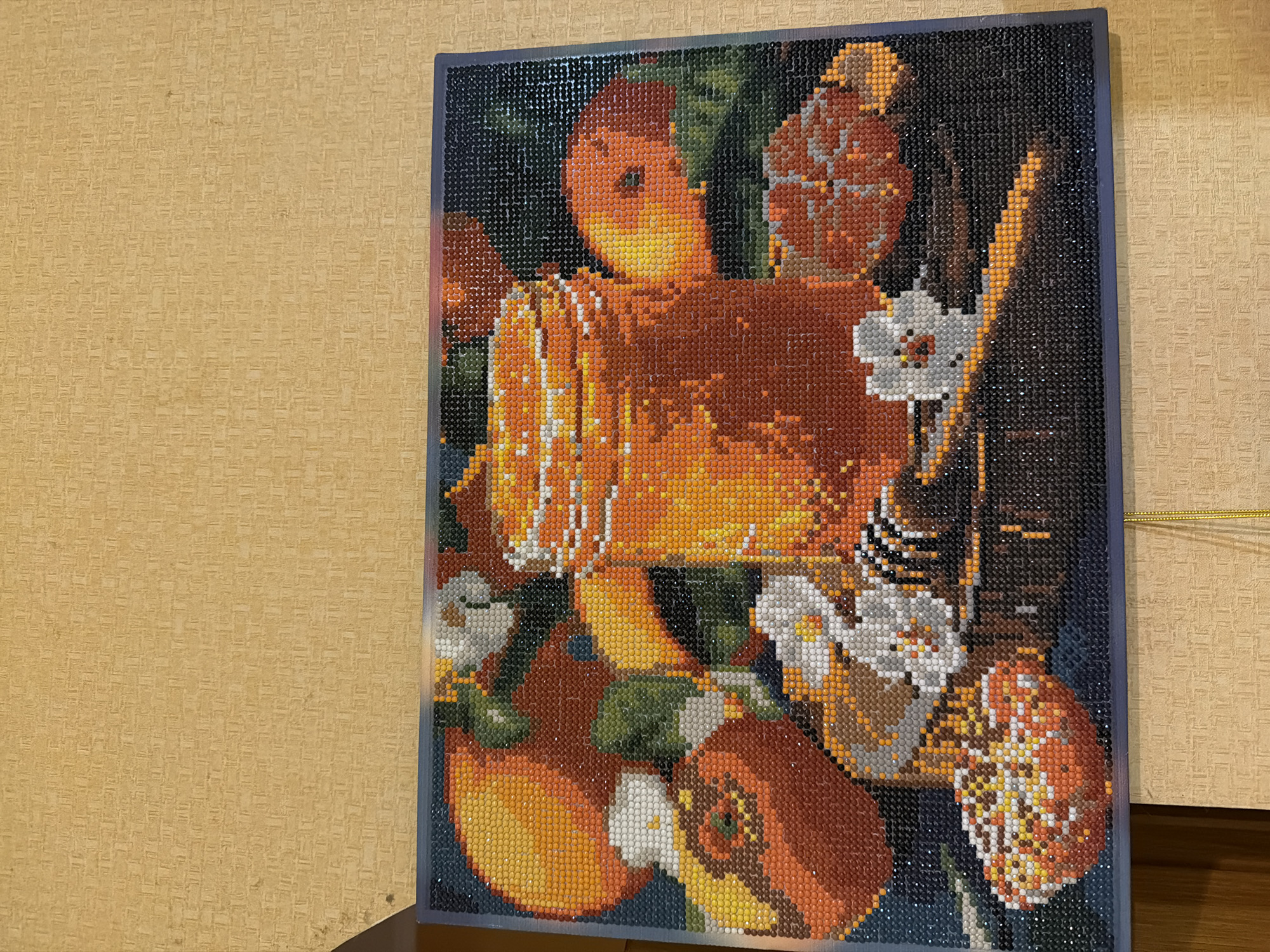 A diamond painting depicts a still life of a glass jar filled with amber-colored honey, placed amongst persimmons and small white flowers. A honey dipper with honey dripping from it lies across the foreground. The background is dark green. The painting is framed and hangs on a textured beige wall. The image appears to be taken indoors, possibly in a home. No specific location in Kyiv, Ukraine can be determined from the image.
