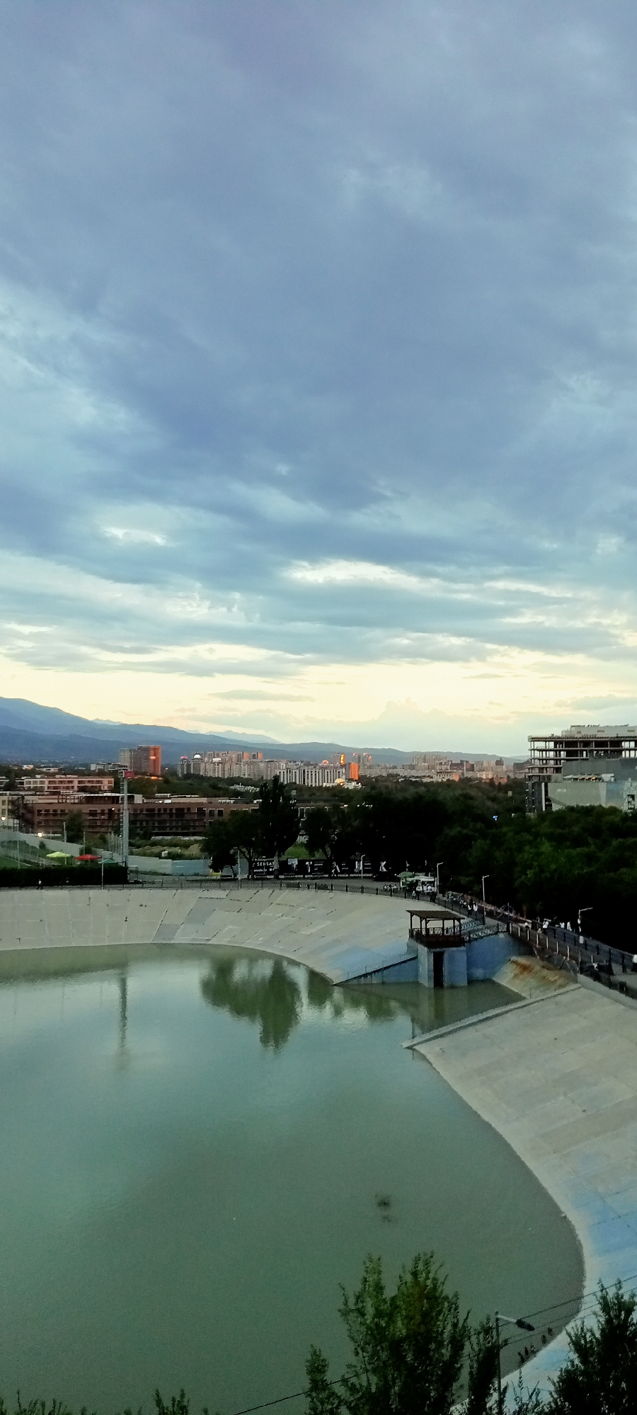 This media file depicts a wide shot of a reservoir and an urban skyline at dusk. The reservoir, with its light green, calm water, occupies the foreground and mid-ground, bordered by a light-colored concrete embankment. A small, elevated structure with a dark roof sits on the embankment, extending over the water. In the background, a dense green treeline separates the reservoir from a city cityscape featuring numerous buildings, some of which are illuminated by the setting sun, casting a warm orange glow. Mountains are visible in the far distance, silhouetted against the twilight sky.
The sky is filled with soft, muted clouds, suggesting the transition from day to night. The overall atmosphere is tranquil and serene. Several people are visible walking along the embankment of the reservoir, and some appear to be on a pedestrian walkway that runs parallel to the water's edge. The scene captures a moment of everyday life in an urban environment as day concludes. No legible text is visible in the image.