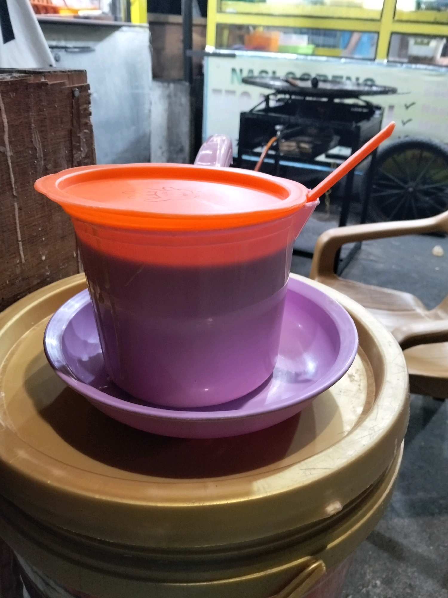 In Arang Limbung, Indonesia, a close-up shot captures a plastic jug with an orange lid and a purple saucer, filled with a dark beverage, likely coffee or a similar drink. The jug is placed on a gold plastic bucket. In the background, a food stall is visible with a stove, cooking equipment, and a sign that reads "NASI GORENG" (fried rice). A wooden structure is to the left of the jug, and a plastic chair is to the right. The lighting suggests it is evening or a dimly lit indoor setting.