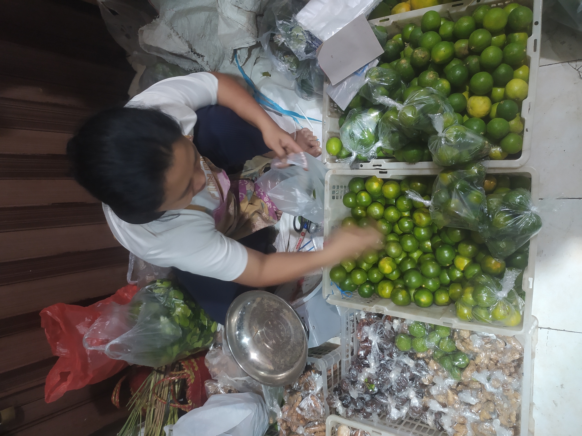 A person is crouched down in what appears to be an outdoor market stall in Kupang, Indonesia, surrounded by produce and goods. The person, with dark hair and wearing a white t-shirt and dark pants, is in the process of bagging some small green fruits, likely limes or calamansi, into a clear plastic bag. In front of them, to their right, are two large white plastic crates overflowing with these small green fruits, some of which are also bagged. To the person's left, there are two more white plastic crates filled with various packaged goods. One crate contains items wrapped in brown paper, possibly dried spices or nuts, while the other holds items in clear plastic packaging, some appearing dark and others lighter brown. A metal weighing scale with a silver bowl is visible on the person's left, positioned between the crates of produce and packaged goods. Behind the person, a red plastic bag and a few other items are partially visible. The background consists of wooden slatted walls and the floor is tiled. The scene suggests a vendor actively engaged in preparing goods for sale in a bustling marketplace environment.
