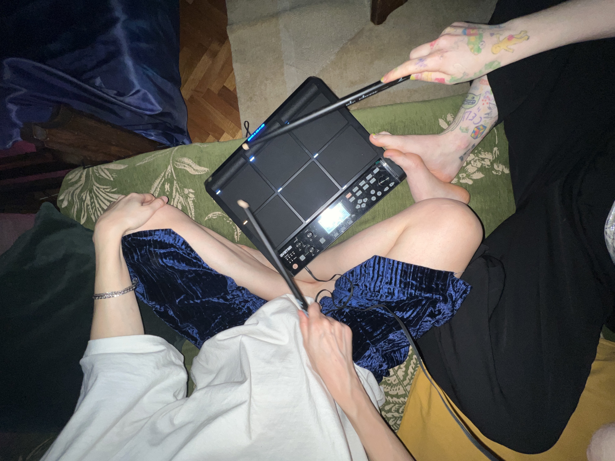 An overhead shot captures an indoor scene where two individuals are engaged with an electronic drum pad. The central focus is a black electronic drum pad, likely a Roland SPD-SX or similar model, resting on the lap of one person. This individual, wearing a white t-shirt and blue velvet shorts, has their legs bent around the device and holds a black drumstick in their right hand, poised to strike the pads. A silver chain bracelet is visible on their left wrist.
Partially visible to the right is a second person, wearing black pants. Their left arm, adorned with numerous colorful, cartoonish tattoos, is holding another black drumstick, also aimed at the drum pad. Their bare feet, also tattooed with similar designs, are visible near the device.
The activity clearly involves playing or practicing music using the electronic drum pad. The pads on the device emit a blue light, indicating it is powered on and possibly in use. The setting appears to be a relaxed, casual indoor space, possibly a living room, with green patterned cushions from a sofa or armchair beneath the people. A dark blue velvet fabric or blanket is draped on the left, and a wooden floor is visible in the background.
Visible text includes "SYSTEM" on the drum pad and "celt. 01 Recording" on one of the drumsticks. The overall ambiance suggests an evening or nighttime activity due to the indoor lighting and glowing device.