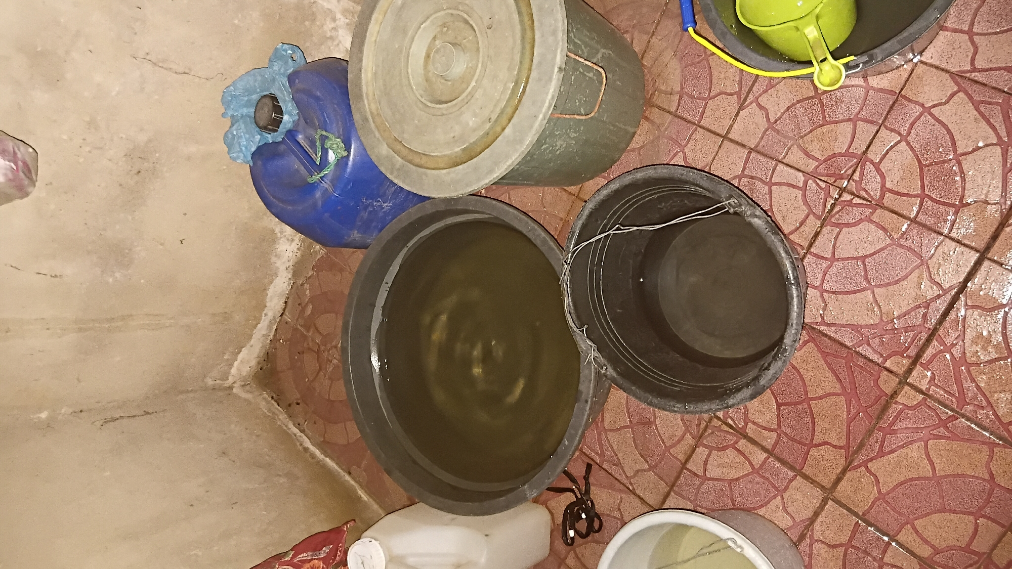 The image depicts a corner of a room with tiled flooring and a textured, off-white wall. The scene is cluttered with various containers, some filled with water. In the center, a large black basin holds murky, yellowish water with visible ripples. To its right, a large blue plastic jerrycan is partially visible, covered with a blue plastic bag secured with green twine. Next to the jerrycan, a large, gray lidded plastic container sits on the floor. To the left of the central basin, a white plastic jug with a white cap is positioned. In the foreground, a smaller black bucket, seemingly empty or containing very little water, has wire handles. Further to the right, a gray basin contains a bright green plastic ladle. Water appears to be spilled or present on the tiled floor, particularly around the containers. The lighting suggests an indoor environment, possibly a storage area or a utility space. The presence of multiple water containers may indicate a need for water storage or collection, potentially due to supply issues or preparation for a task. The scene appears to be located within a residential or communal setting in Prabumulih, Indonesia, given the context provided.