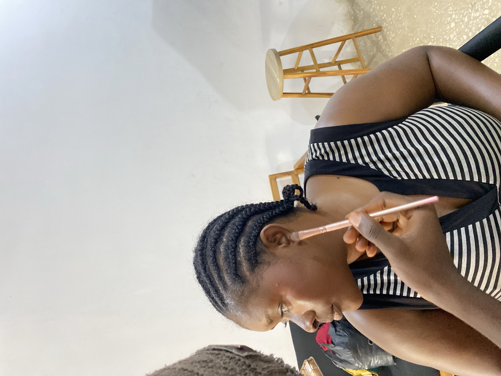 The photograph depicts a woman receiving makeup application. Her hair is styled in braids, and she is wearing a black and white striped top. A makeup brush is being held near her face, presumably to apply makeup. In the background, a wooden stool is visible. The location is not specified.