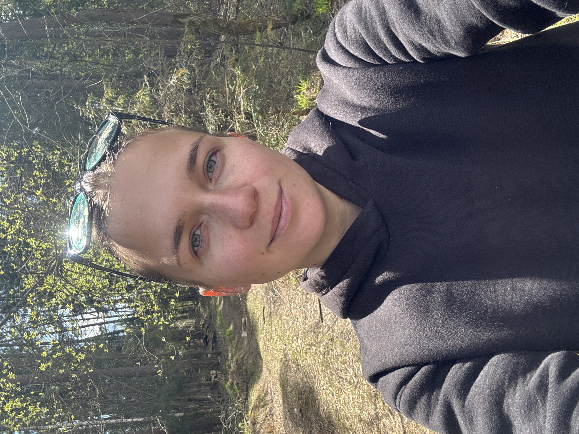 A woman is photographed outside in Раздолье, Russia. She wears a dark hooded sweatshirt, with sunglasses pushed up on her head. The location is a wooded area with trees in the background and vegetation and a path visible behind her. The lighting suggests it is a sunny day.