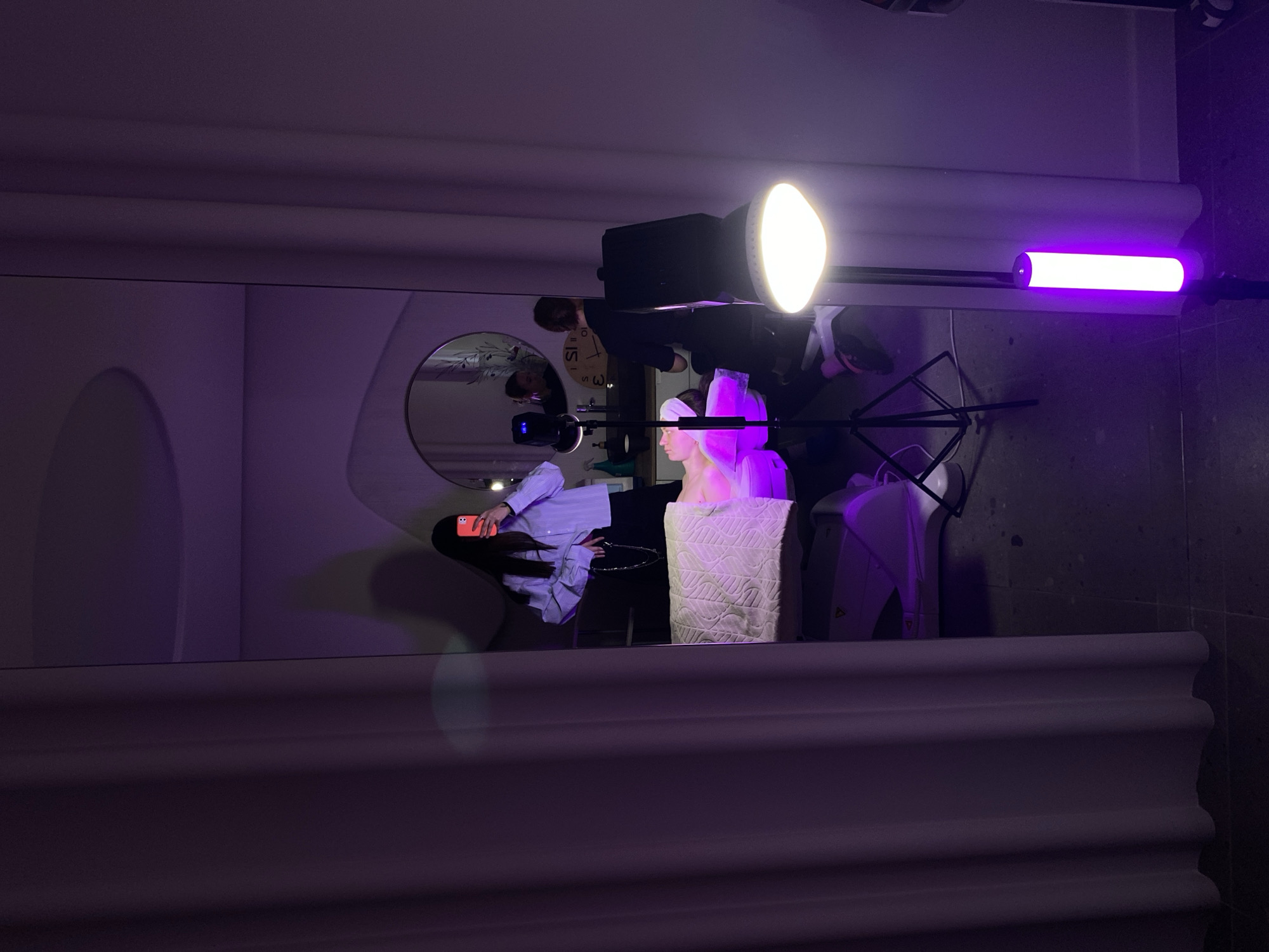 The image shows a mirrored view of a room with studio lighting setup. Reflected in the mirror is a person holding a mobile phone, wearing a light blue collared shirt. To the right of this figure is a white object with textures, potentially a pillow or cushion. A studio light with a reflector is aimed at the object. To the right, a purple light stick is visible. In the background of the mirrored image, there is a round mirror on the wall and what appears to be a person standing near a wall-mounted clock. The location is unknown.