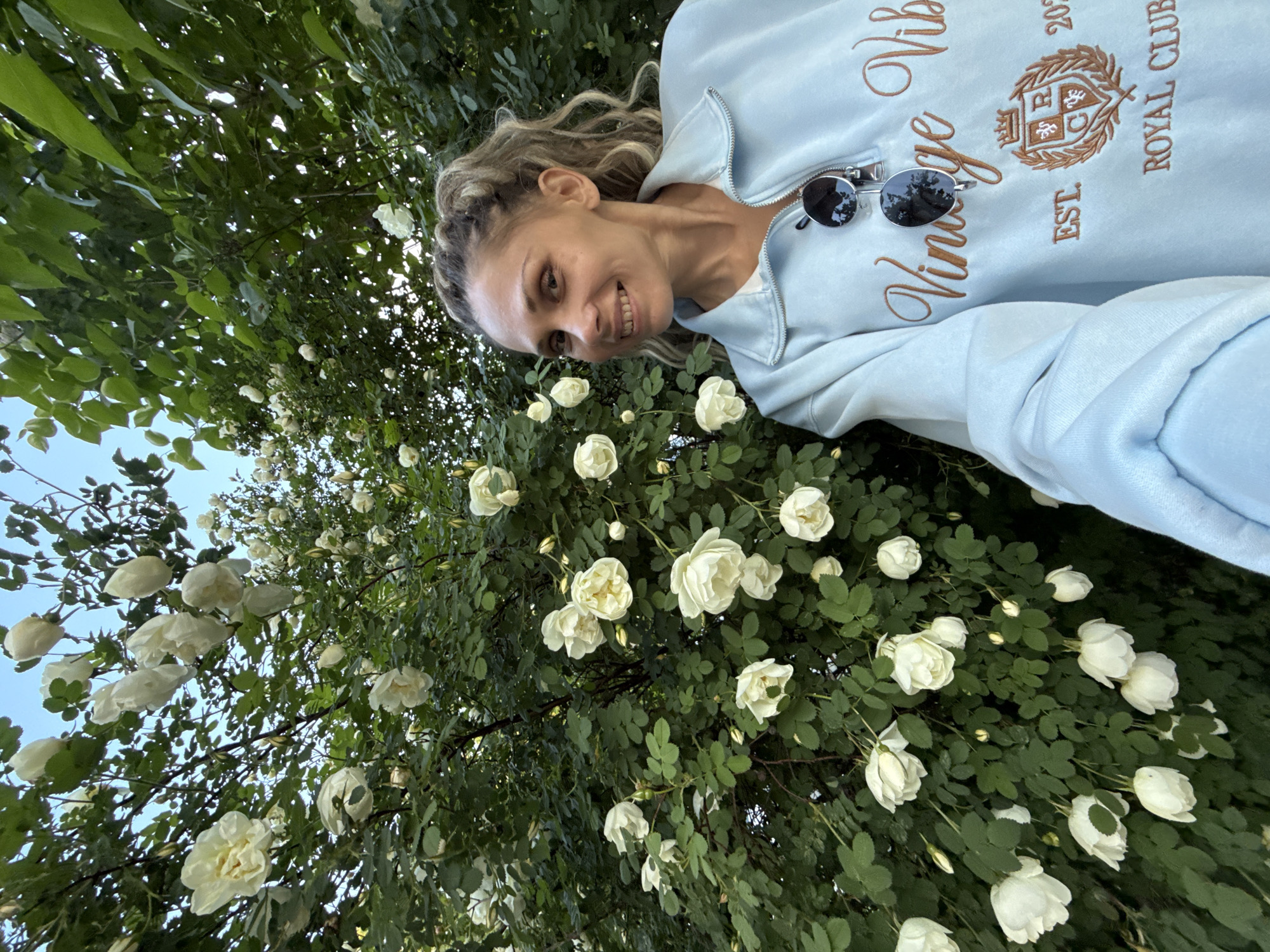 A woman with blonde hair is posing for a selfie in front of a flowering bush with white roses. She is wearing a light blue sweatshirt with the words "Vintage Vibes Royal Club EST 2023" embroidered on it and sunglasses hanging from the collar. The bush is densely covered with white roses and green leaves. The sky is visible in the upper left corner. The location is Rusanovo, Russia.