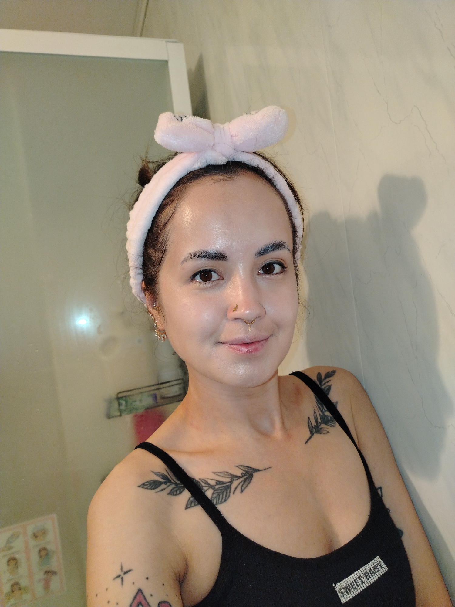 A young woman is captured in a waist-up selfie, smiling softly at the camera from an indoor setting in Noviy, Russia. She has dark hair pulled back and wears a light pink, fluffy headband with a bow detail at the top. Her complexion is clear and dewy, and she has dark eyes. She sports a gold septum piercing with a dangling bead and a gold stud in her right nostril. Her left ear features elaborate, dangling gold earrings.
She is wearing a black tank top with the white text "SWEET BABY" printed on the chest. Several tattoos are visible: a leafy branch design adorns her left collarbone and shoulder, another similar branch extends across her right chest, and geometric shapes, including a star and a diamond, are partially visible on her right upper arm.
The background suggests a bathroom or similar room, with light-colored, possibly beige, tiled walls featuring subtle patterns. To her left, a reflective surface, likely a mirror or shower door with a white frame, reflects blurred light, hinting at artificial indoor lighting. The overall impression is a relaxed and personal moment.