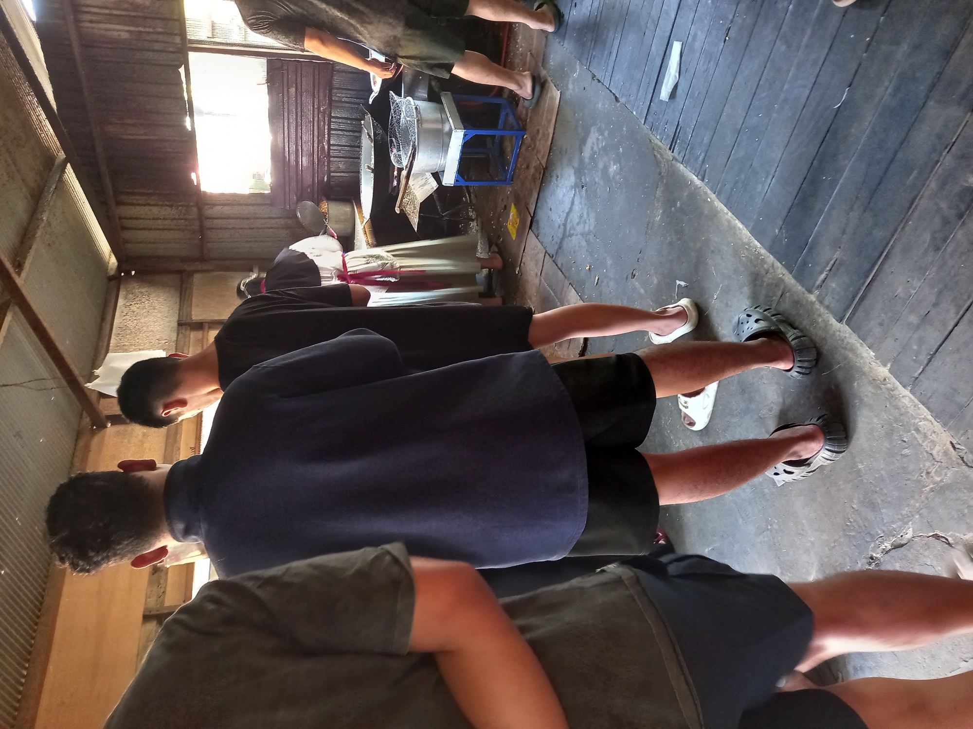 The image depicts an outdoor food stall in Samarinda, Indonesia, likely a temporary setup. The foreground features the legs of several individuals, all wearing casual attire such as shorts and flip-flops or clogs. A man in the center is wearing a dark blue t-shirt and black shorts. To his left, another person wears a dark grey t-shirt and dark shorts. To the right, a third person is seen wearing a dark grey t-shirt and dark shorts, with their legs extended forward. In the background, a person in a white and red garment appears to be cooking. The environment consists of a corrugated metal roof supported by wooden beams, and walls made of corrugated metal sheets. The floor is a mix of concrete and wooden planks. Cooking equipment, including pots and a steamer basket, is visible on a blue-legged stand. The overall impression is a bustling, informal food service area.