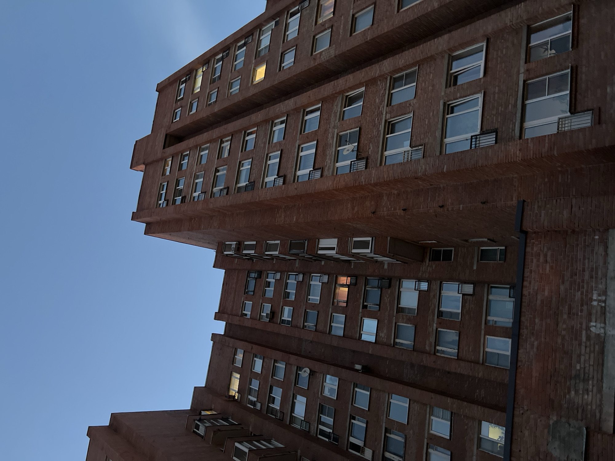 This media file is an upward-looking shot of a Brutalist-style apartment building, possibly in Saint Petersburg, Russia. The building's exterior is constructed from dark red brick and features numerous windows. Some windows are lit from within, suggesting that it is evening or night. The sky is a clear, pale blue, indicating fair weather. The architecture is characterized by its imposing, angular form and repetitive window patterns. There is no visible text in the image. The overall impression is one of urban, residential architecture captured at a specific time of day.