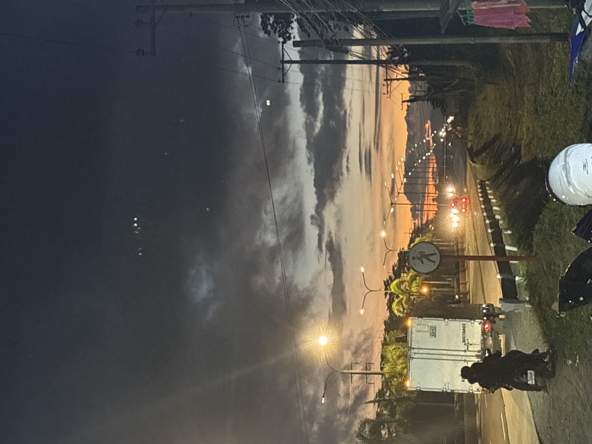 The image depicts a roadside scene at dusk in Carmen, Philippines. The sky is a dramatic mix of dark, cloudy upper portions and a vibrant orange and yellow sunset near the horizon. Several streetlights line the road, some illuminated, creating a warm glow against the twilight. On the left, a white truck with "DISTANCE" visible on its side is parked or moving slowly. In front of the truck, two figures are on a motorcycle, one driving and the other as a passenger, both appearing to be wearing dark clothing. A pedestrian crossing sign with a silhouette of a person walking is visible on the right side of the road. The right side of the image shows a grassy embankment with power lines and poles. A few pieces of clothing, a pink and a white garment, are hanging on a line near the power pole. The foreground shows parts of two motorcycles, one with a white helmet resting on it. The road appears to be a multi-lane highway with other vehicles visible in the distance, their headlights and taillights creating streaks of light. The overall atmosphere is that of a busy road as daylight fades, with elements of both urban infrastructure and natural beauty in the sky.