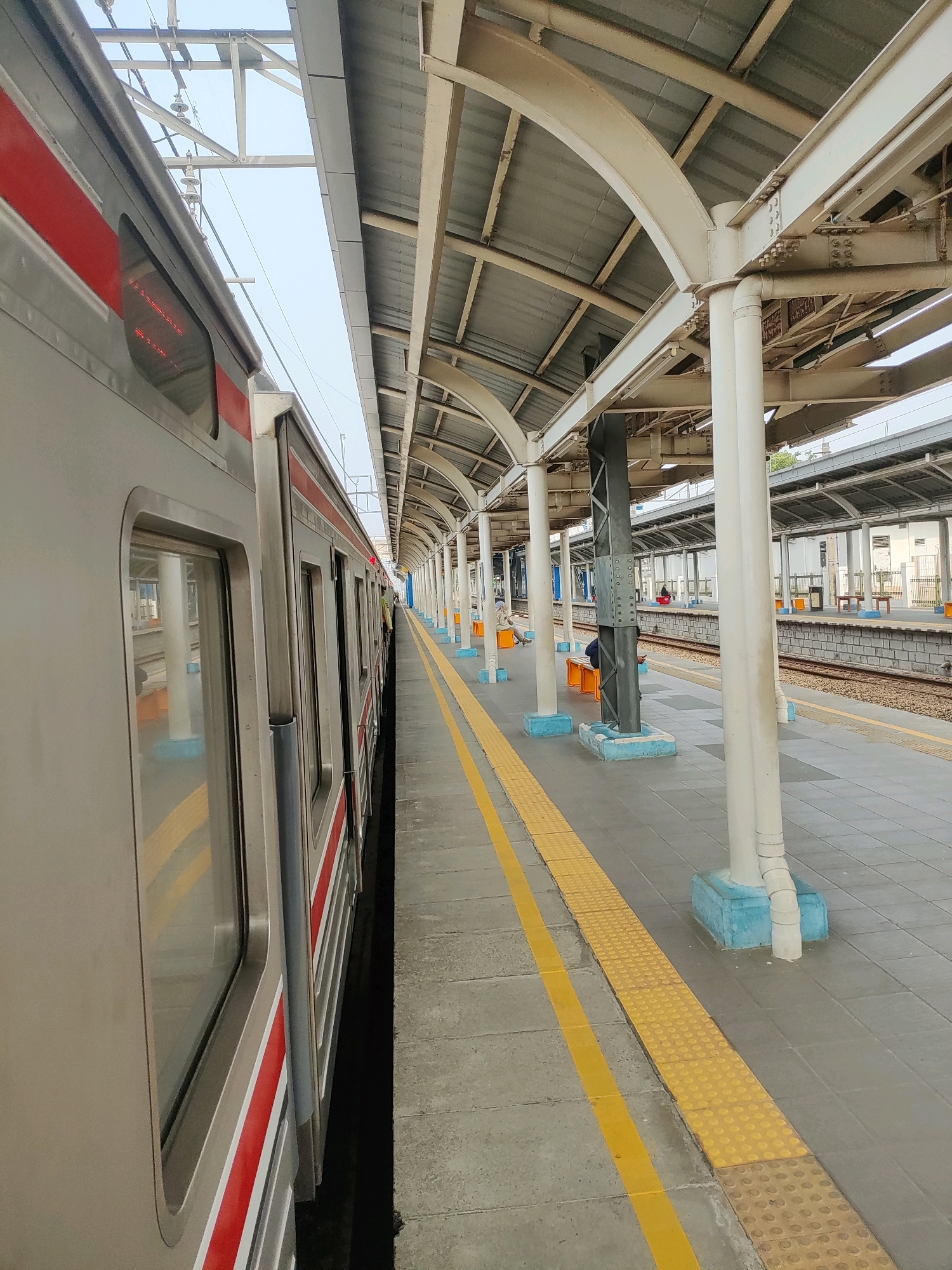 The image shows a train parked at a station platform. The train is silver with red stripes. The platform is paved with grey tiles and has yellow safety lines along the edge. There are white support columns and a metal roof structure. In the background, there are train tracks and other platforms. A few people are visible sitting on benches on the platform. The scene appears to be in Tambun, Indonesia, likely a train station.